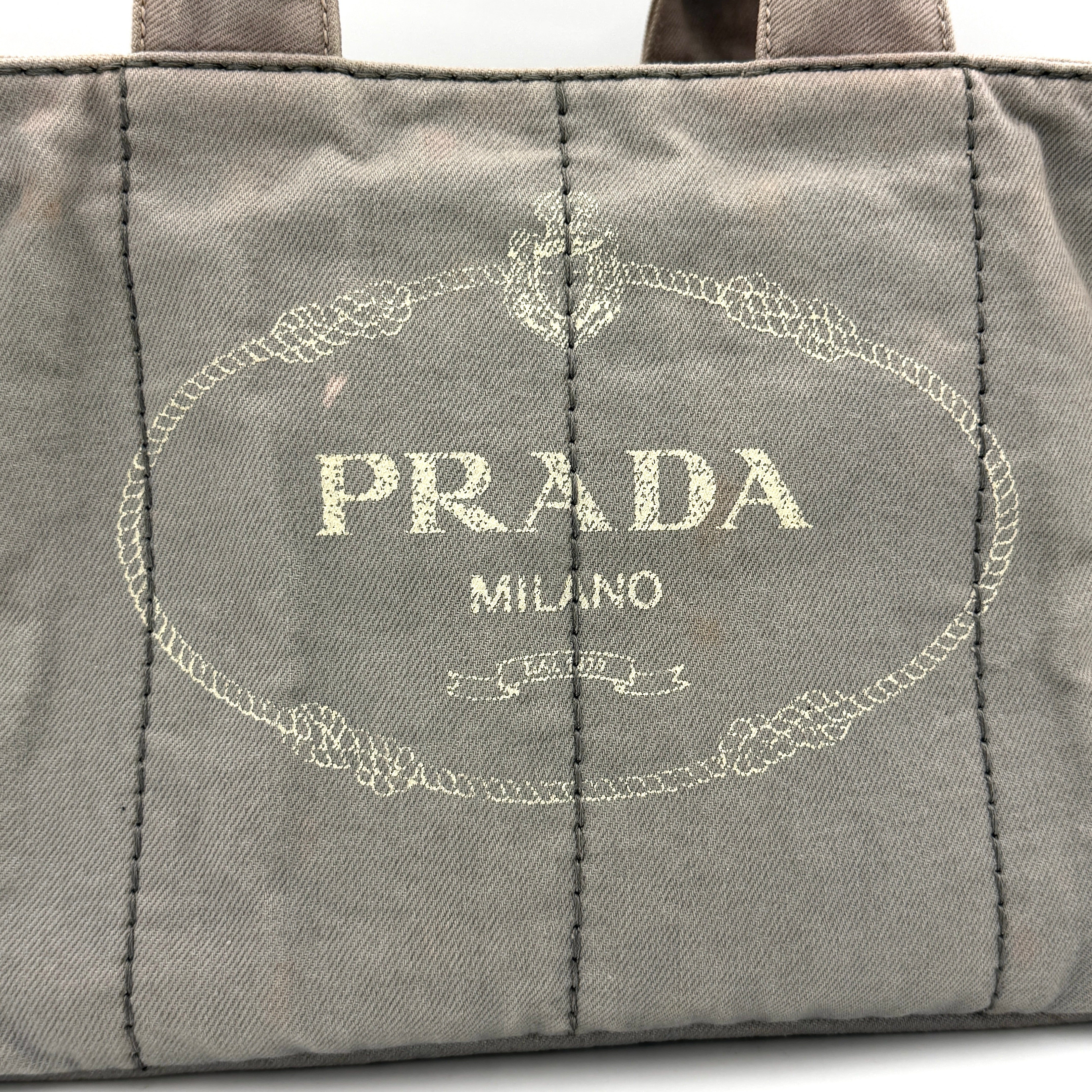 Prada Canapa Tote Bag in grey canvas with rope logo motif and dual top handles against a plain background.