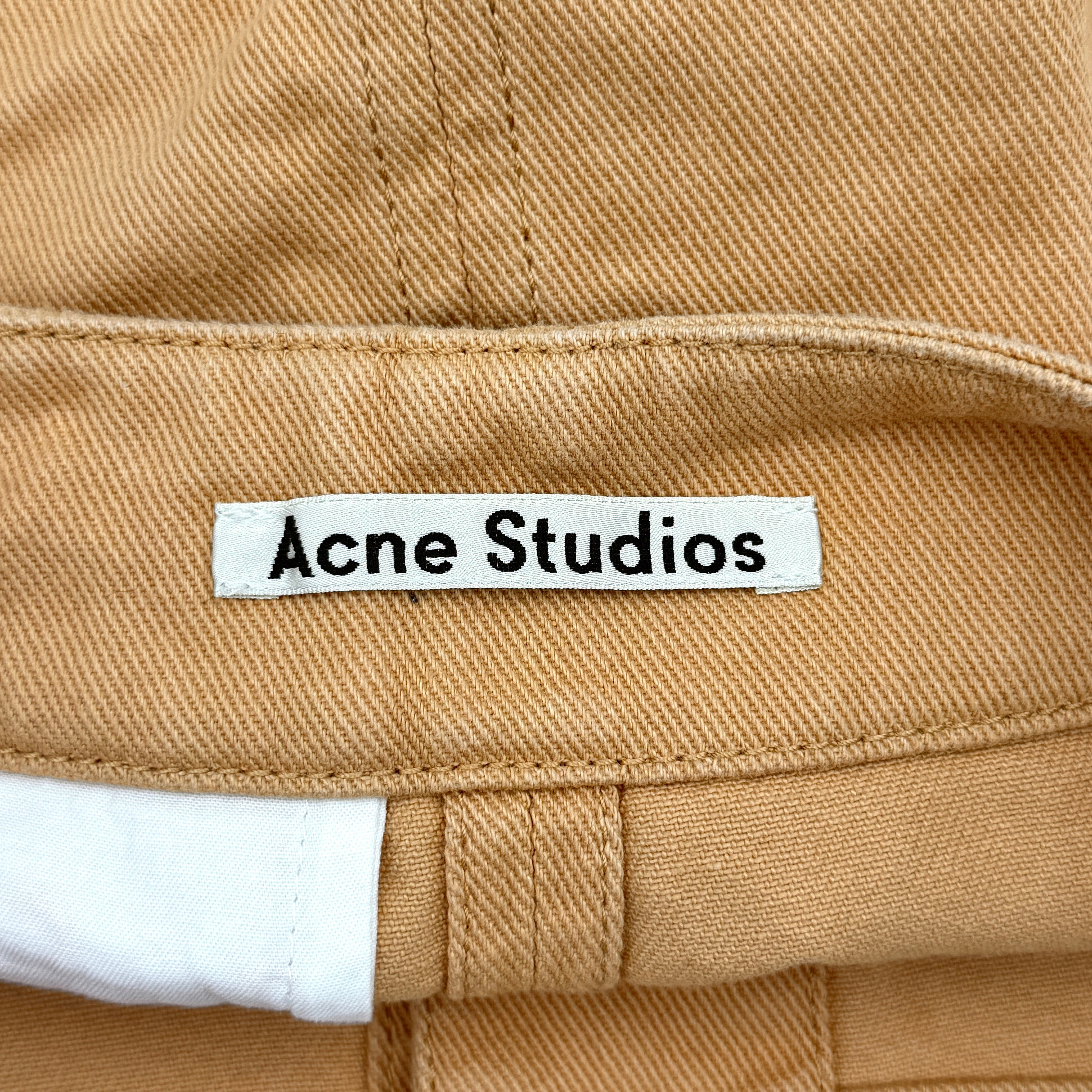 Acne Studios cream and tan cotton drill mini skirt with oversized flap pockets and front button closure, styled flat.