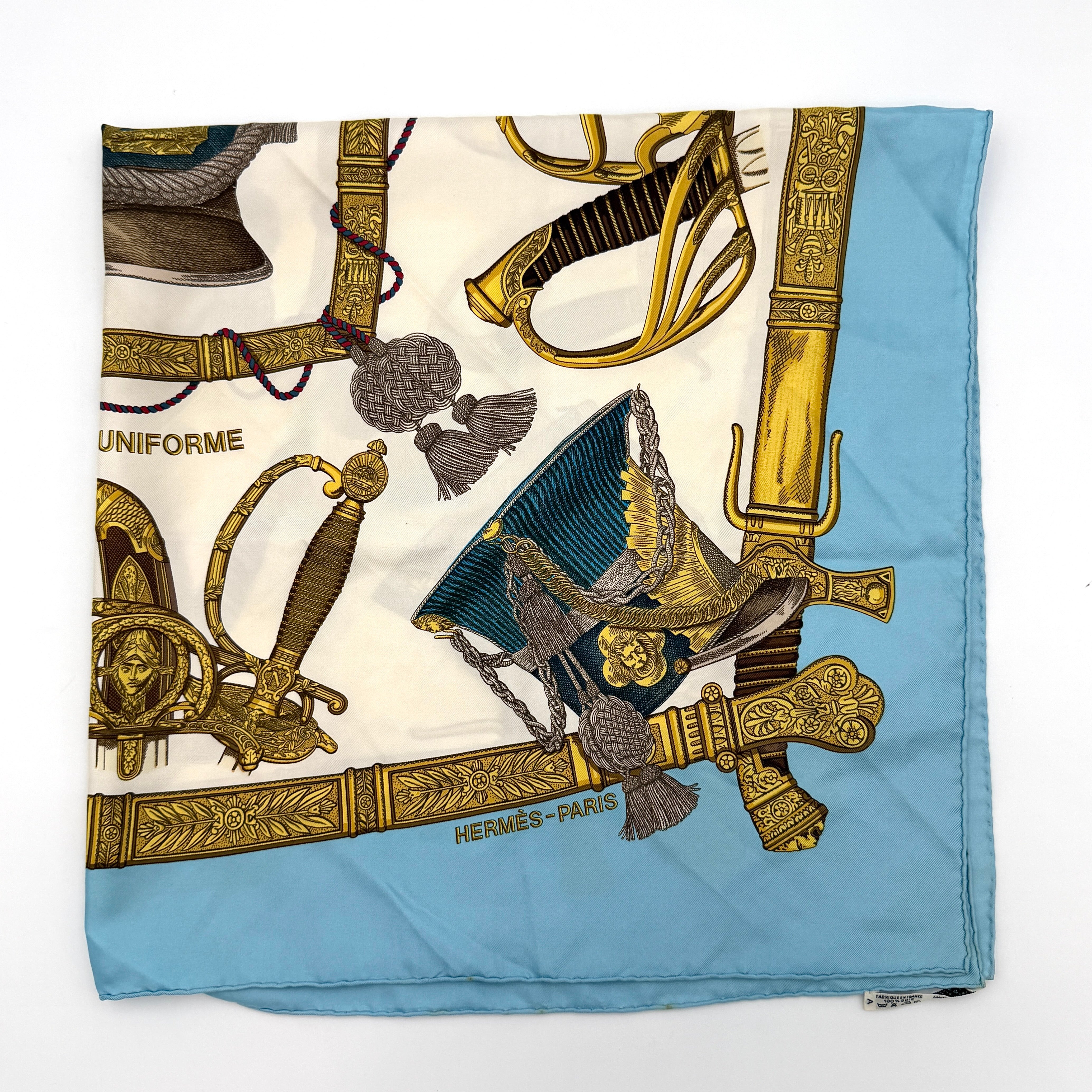 Hermès Grand Uniforme silk scarf with intricate gold and blue Napoleonic-inspired motifs framed on a soft blue and ivory background.