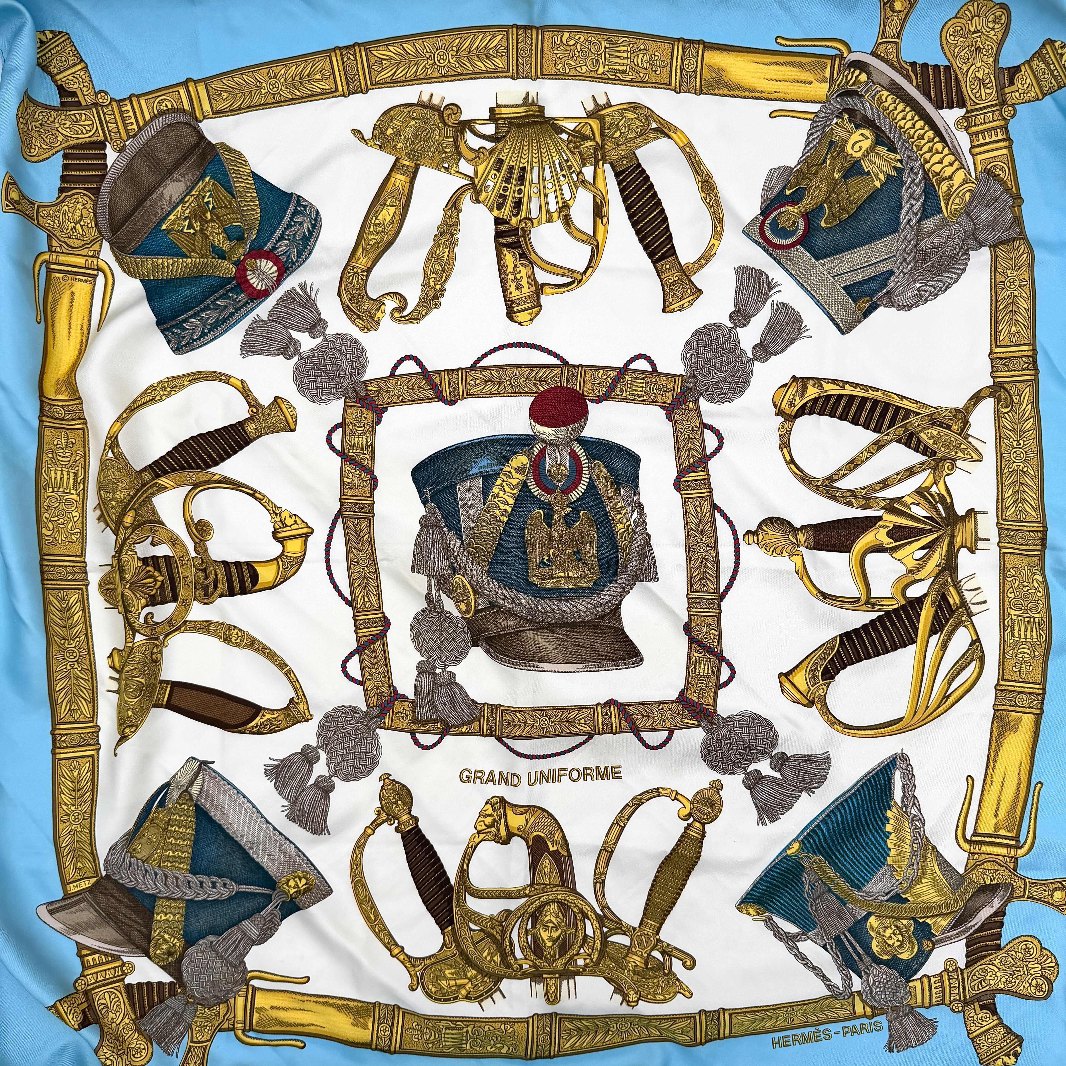 Hermès Grand Uniforme silk scarf with intricate gold and blue Napoleonic-inspired motifs framed on a soft blue and ivory background.