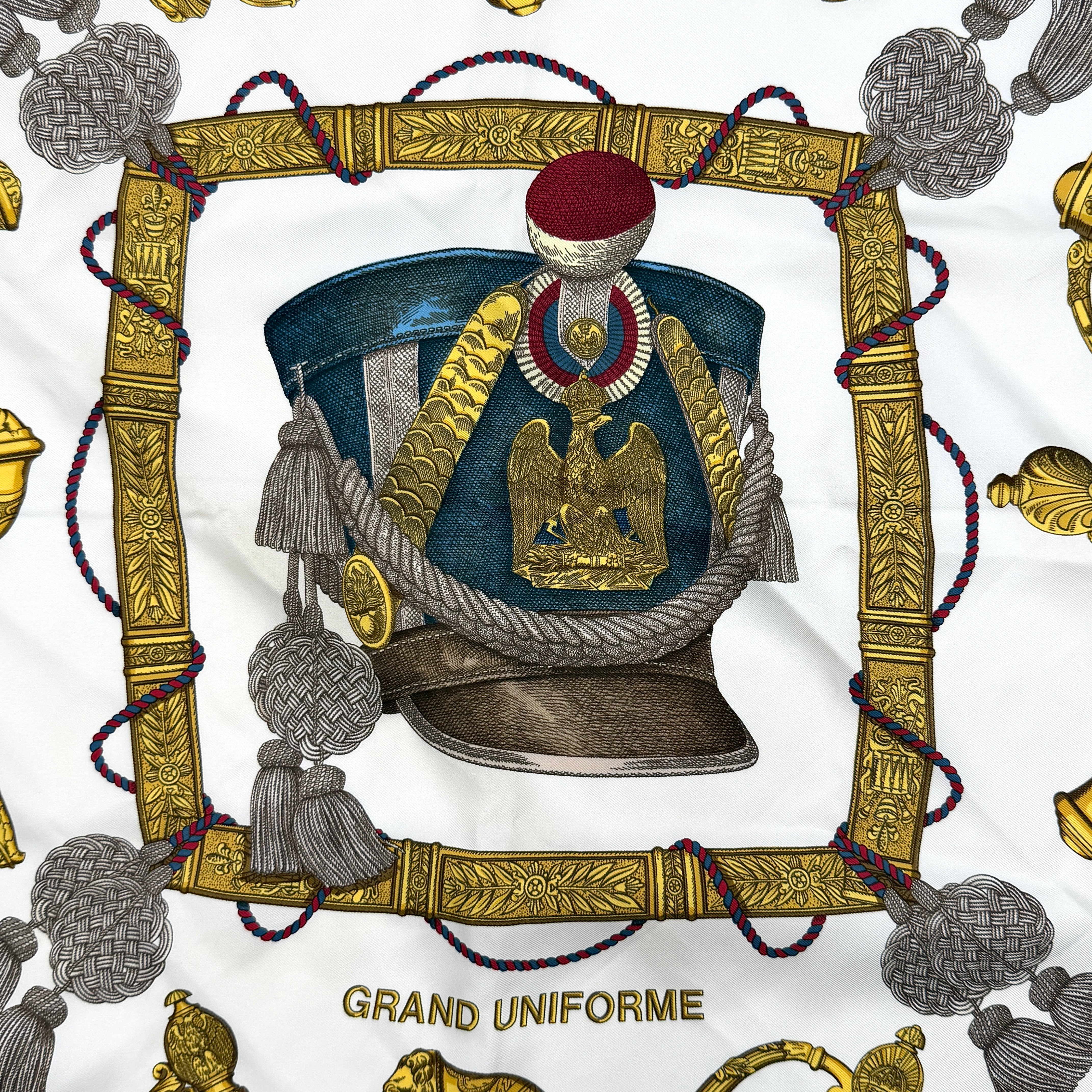 Hermès Grand Uniforme silk scarf with intricate gold and blue Napoleonic-inspired motifs framed on a soft blue and ivory background.