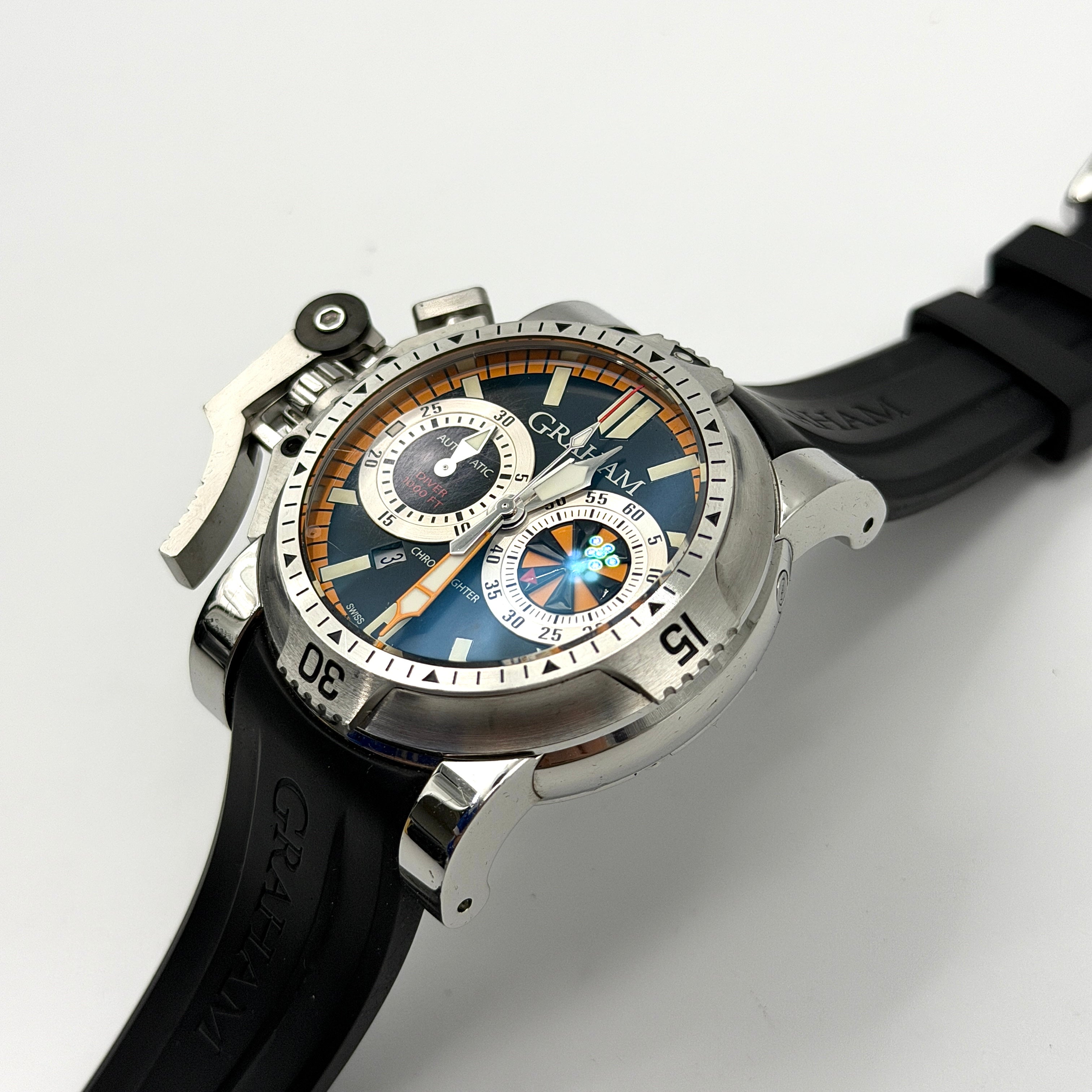 The Graham Chronofighter Oversize Diver 2OVES.B15A watch displayed in its signature round Graham London presentation case, showcasing its bold black dial, prominent chronograph trigger, and black rubber strap.