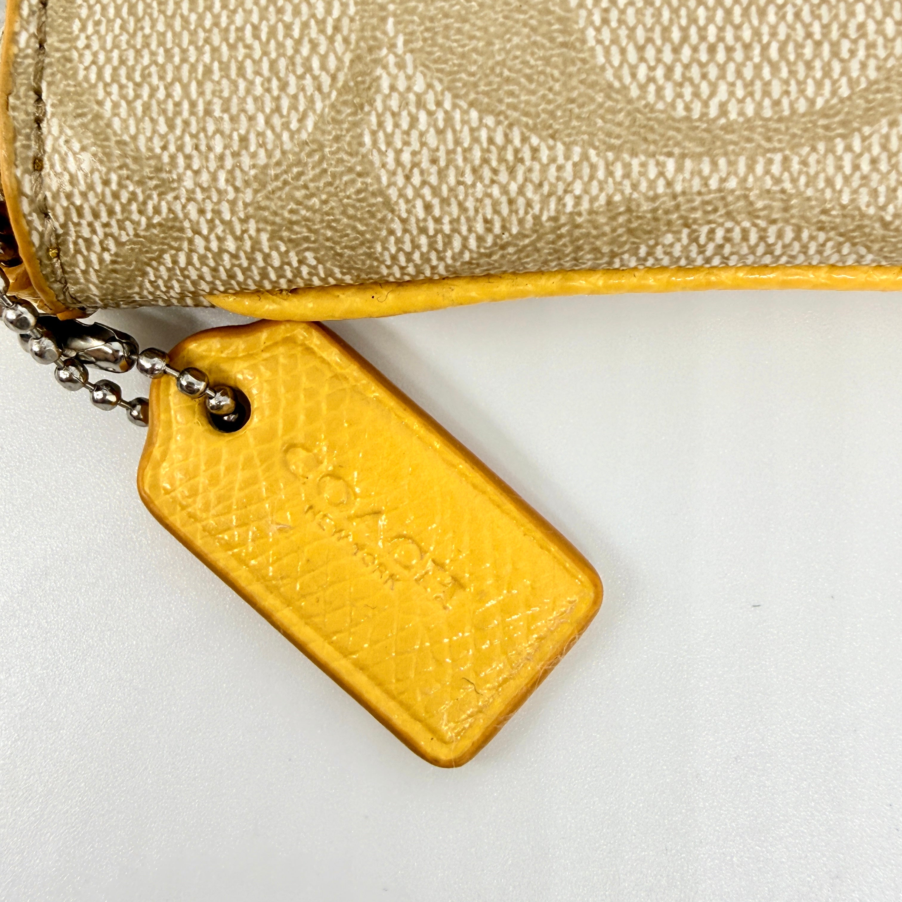 Coach Signature Stripe Corner Zip Wristlet in beige canvas with bold yellow leather stripe, silver hardware, and wrist strap displayed on a white background.
