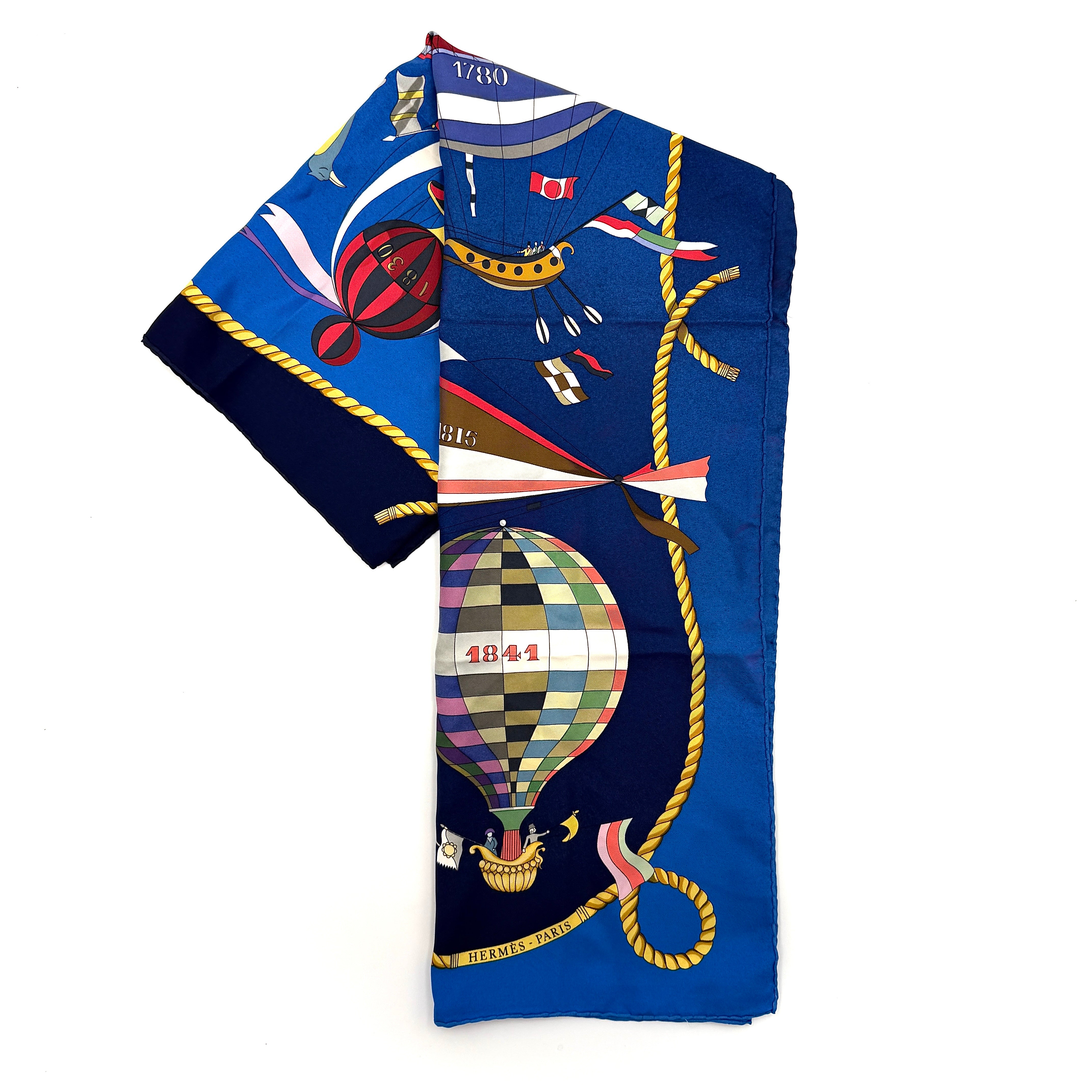 Hermès Les Folies du Ciel silk scarf in vivid multicolours with whimsical hot air balloons and golden rope border on a blue background.