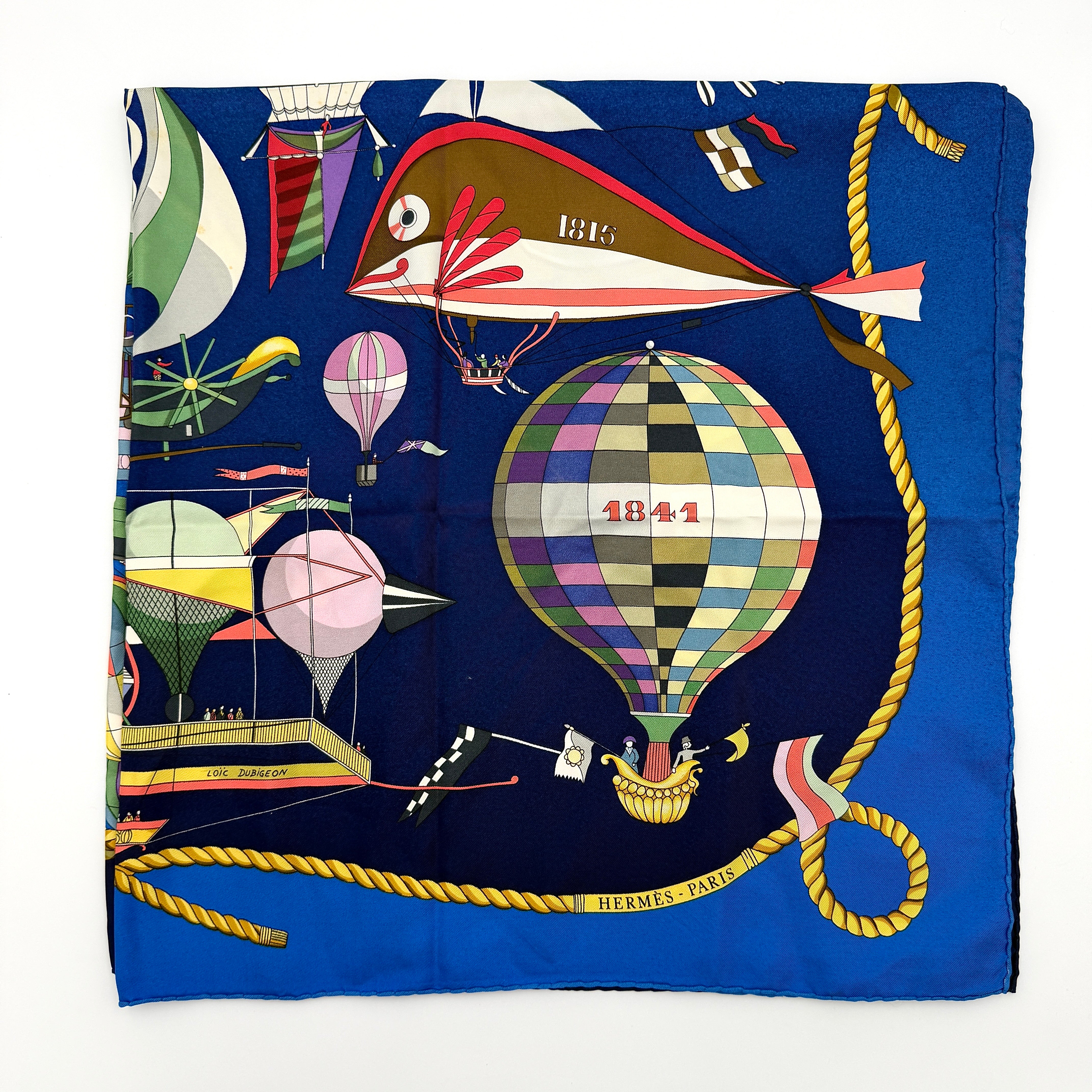 Hermès Les Folies du Ciel silk scarf in vivid multicolours with whimsical hot air balloons and golden rope border on a blue background.