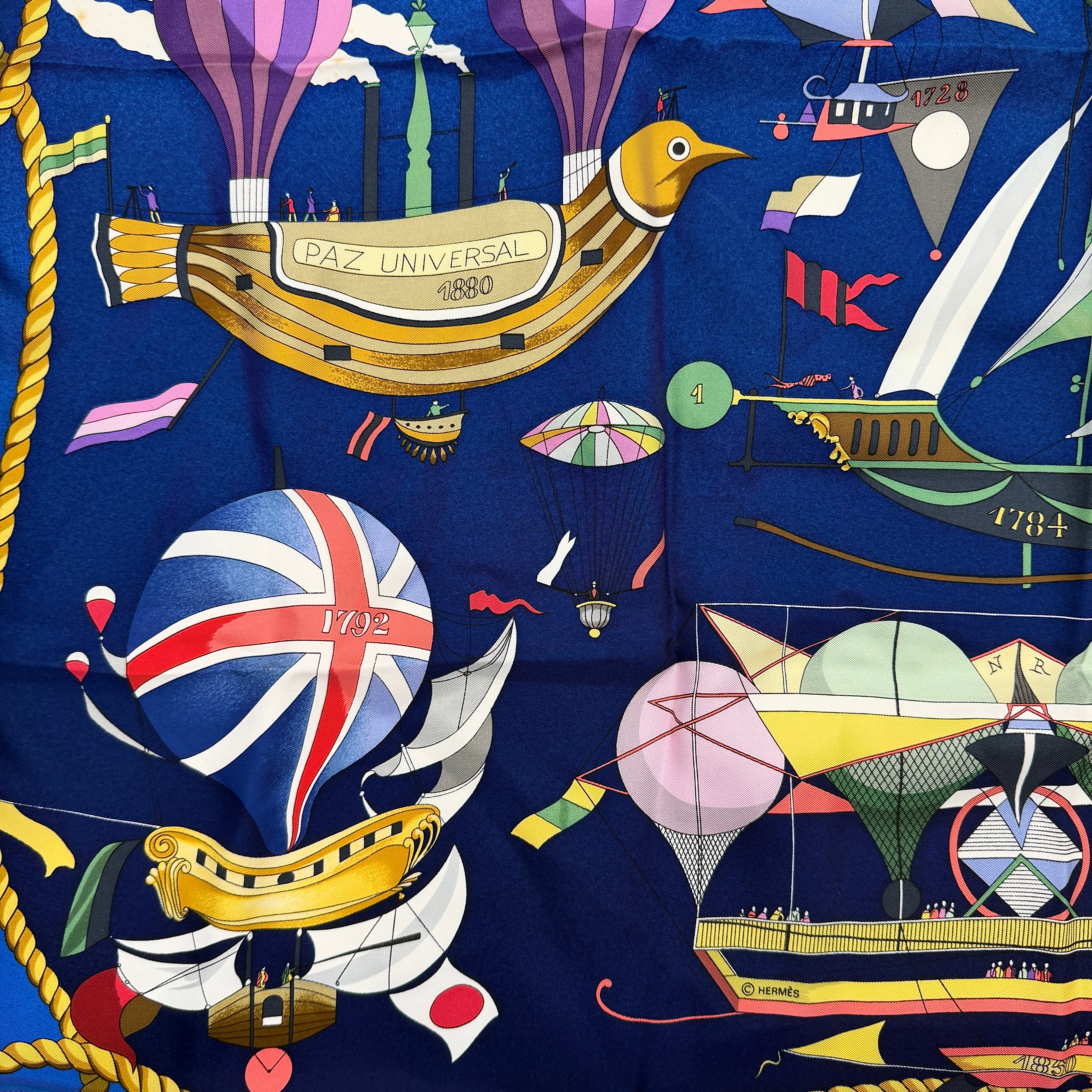 Hermès Les Folies du Ciel silk scarf in vivid multicolours with whimsical hot air balloons and golden rope border on a blue background.