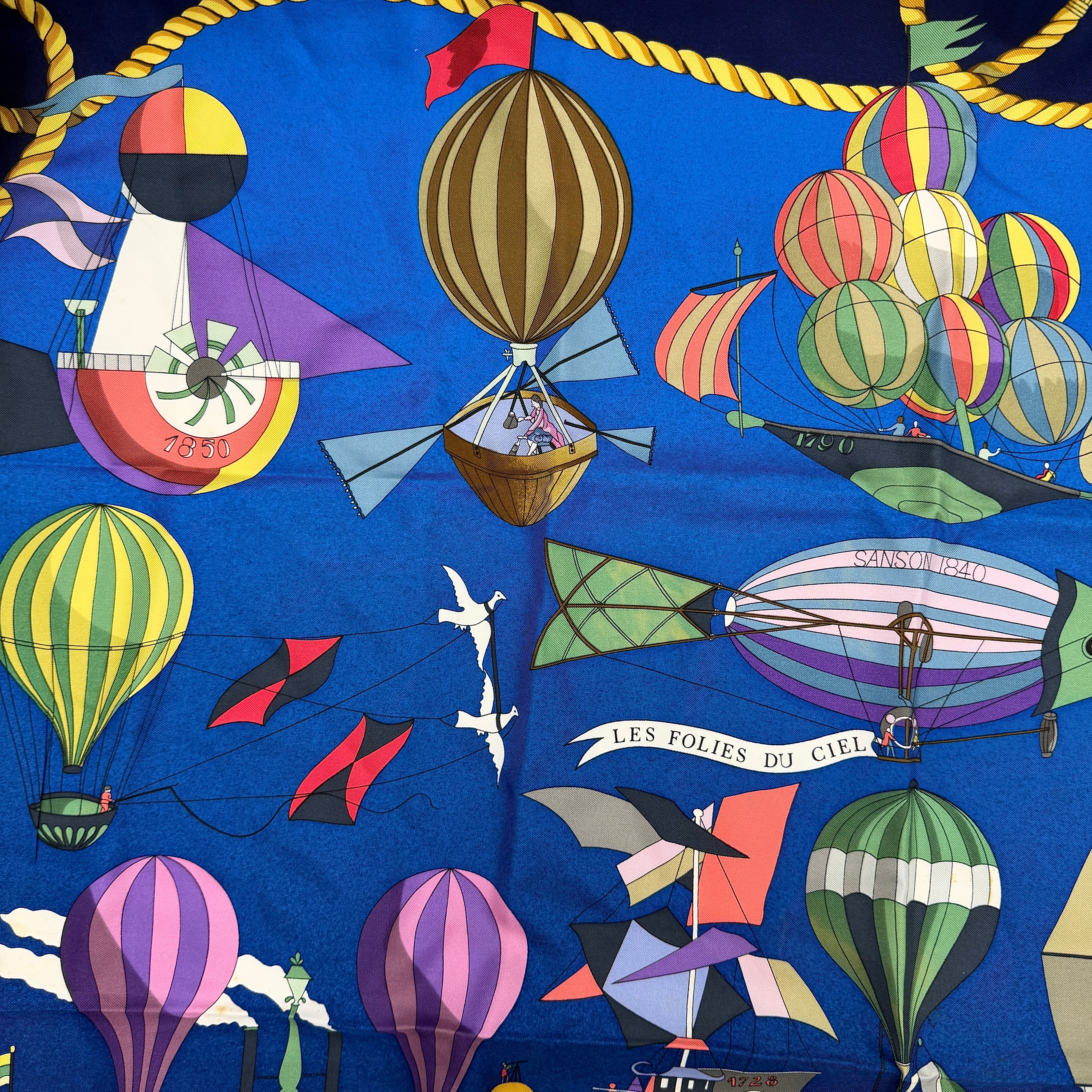 Hermès Les Folies du Ciel silk scarf in vivid multicolours with whimsical hot air balloons and golden rope border on a blue background.