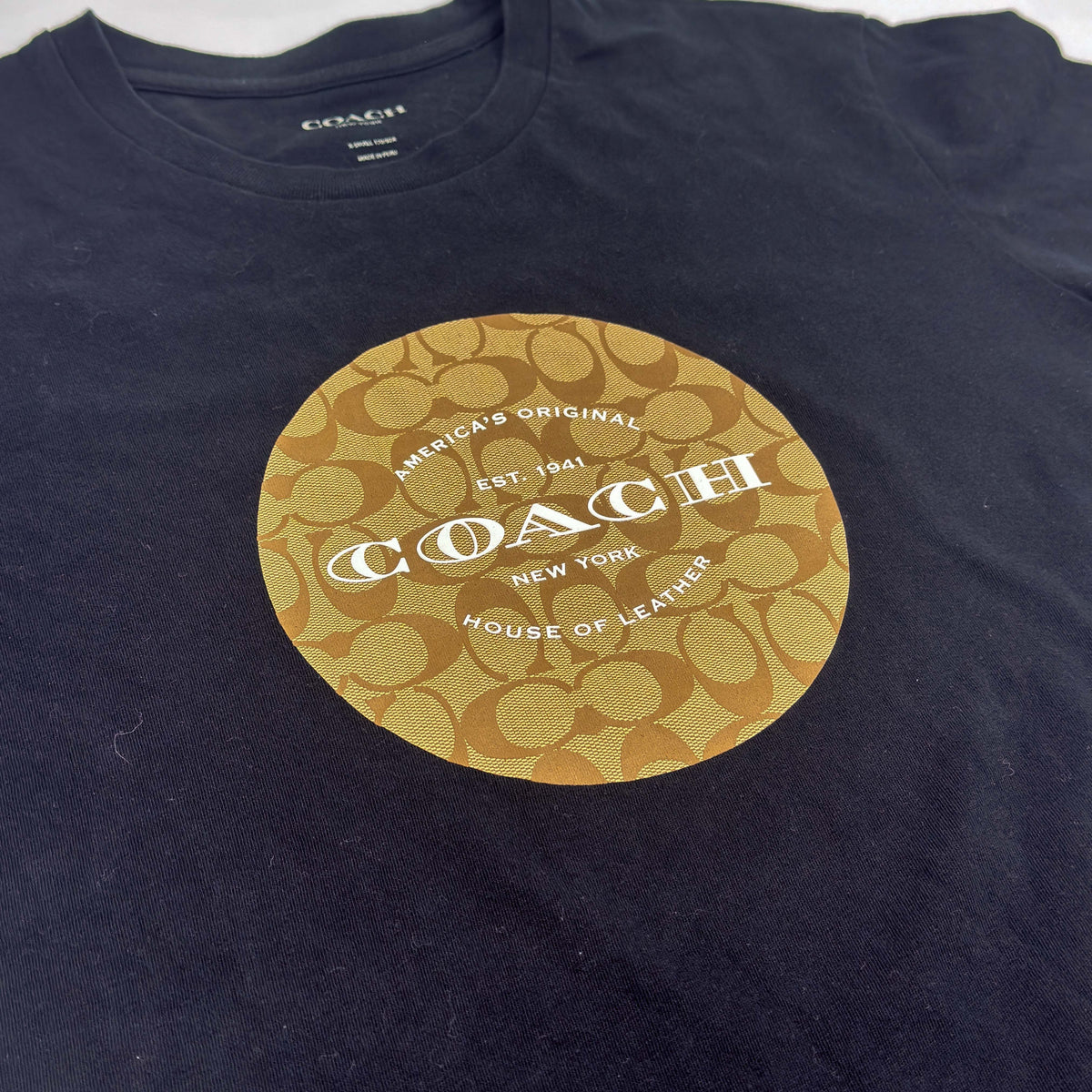 Coach black t-shirt featuring a beige signature logo print on the chest, displayed flat against a white background.