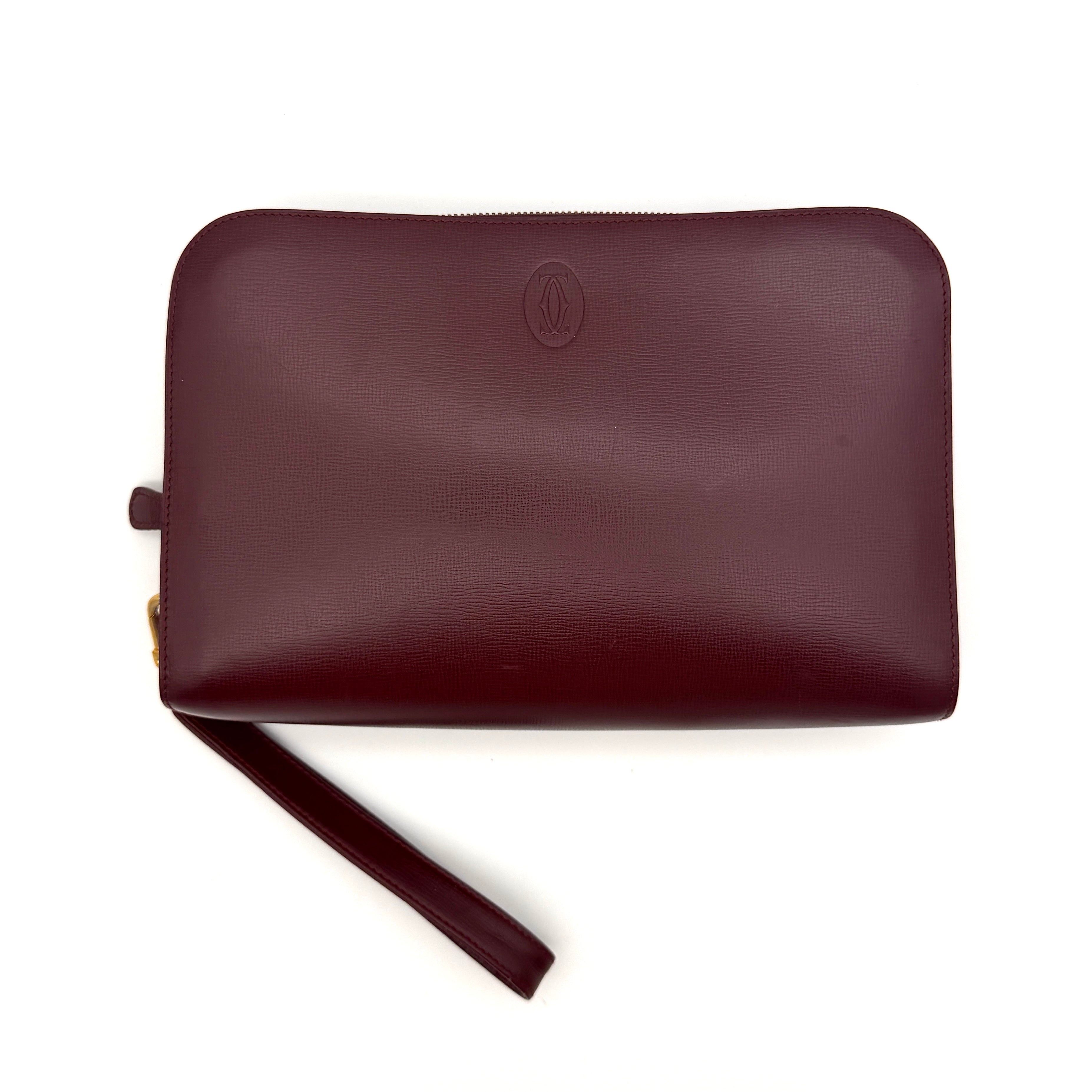 Cartier Must de Cartier clutch in burgundy leather with gold-tone zip and wrist strap, shown against a white background.