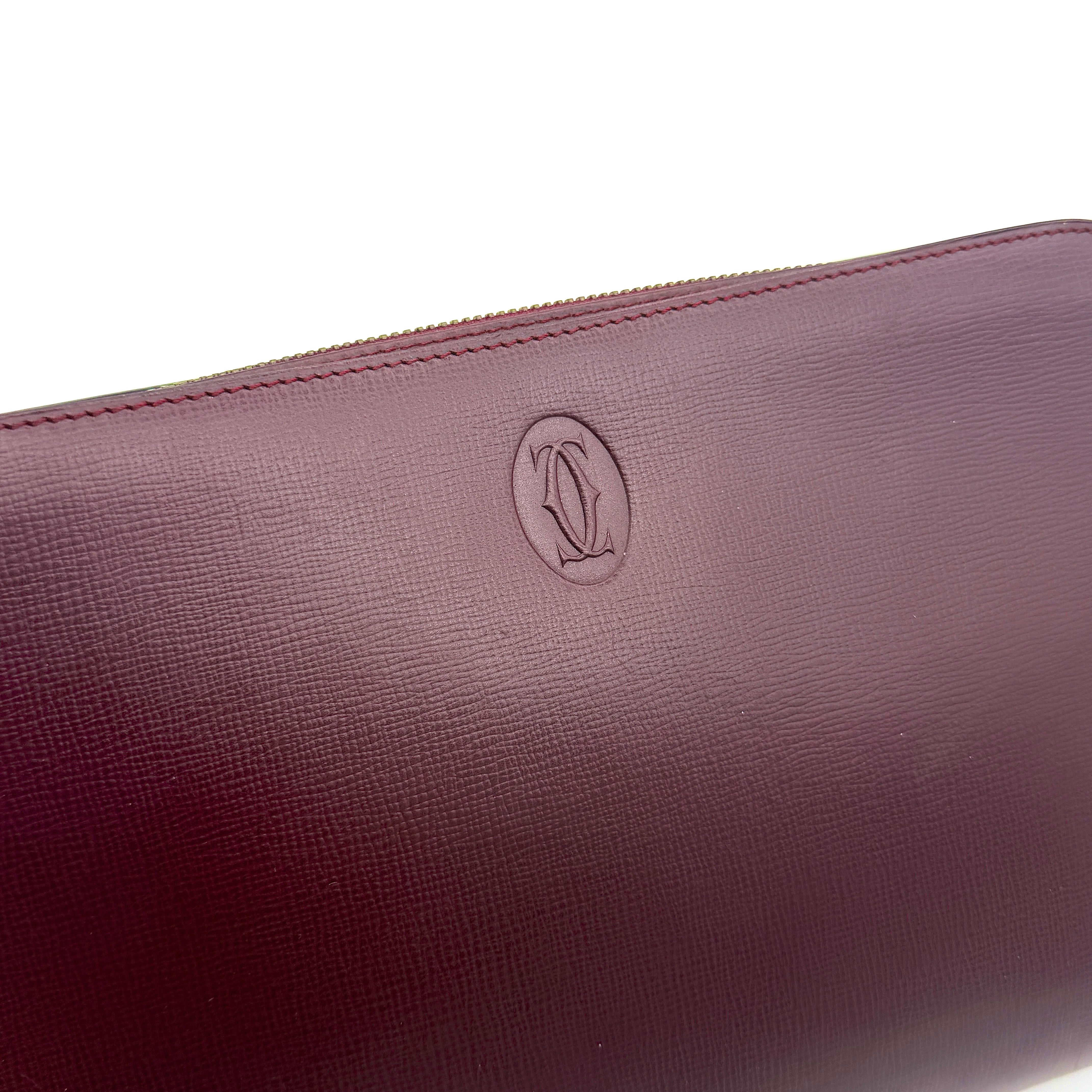 Cartier Must de Cartier clutch in burgundy leather with gold-tone zip and wrist strap, shown against a white background.