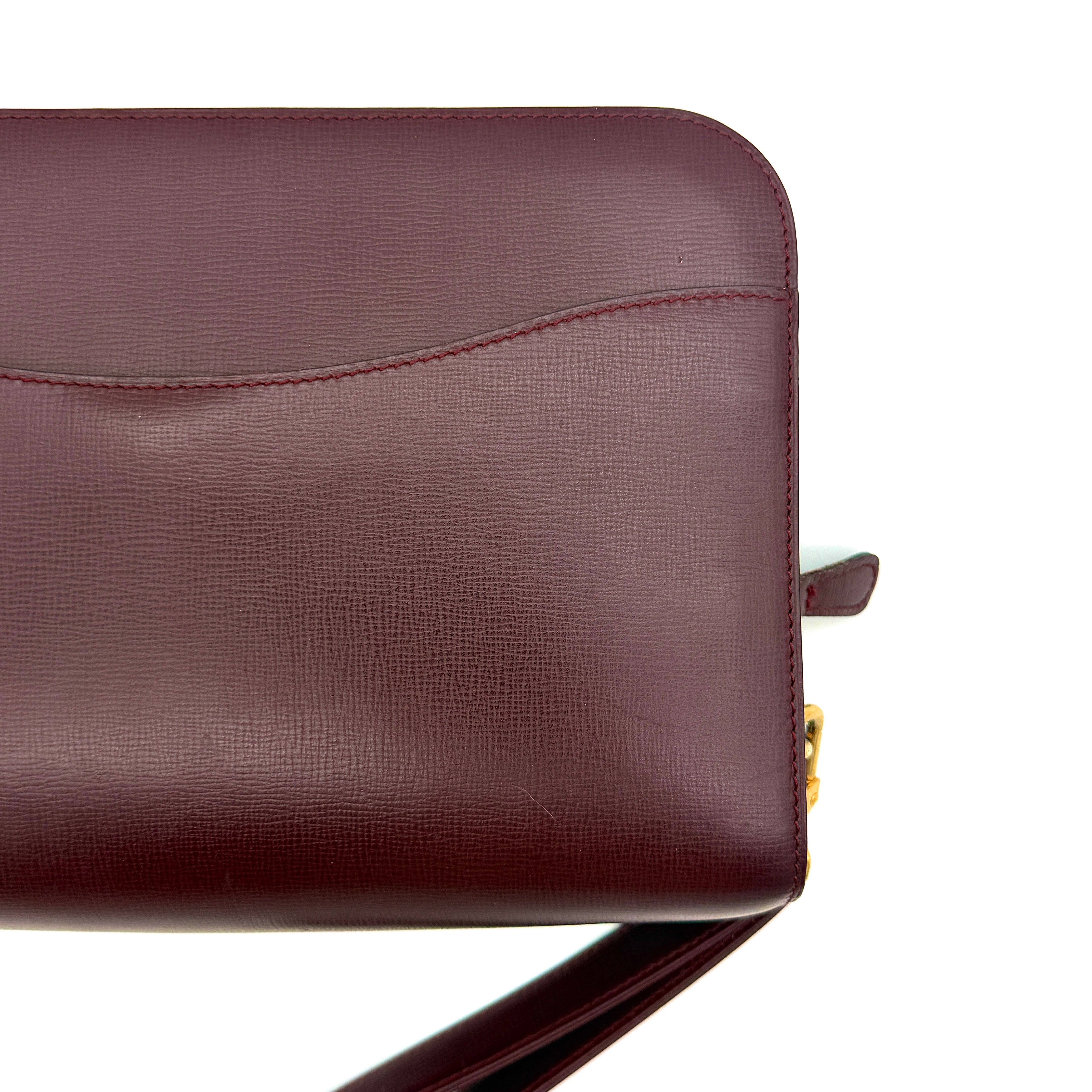 Cartier Must de Cartier clutch in burgundy leather with gold-tone zip and wrist strap, shown against a white background.