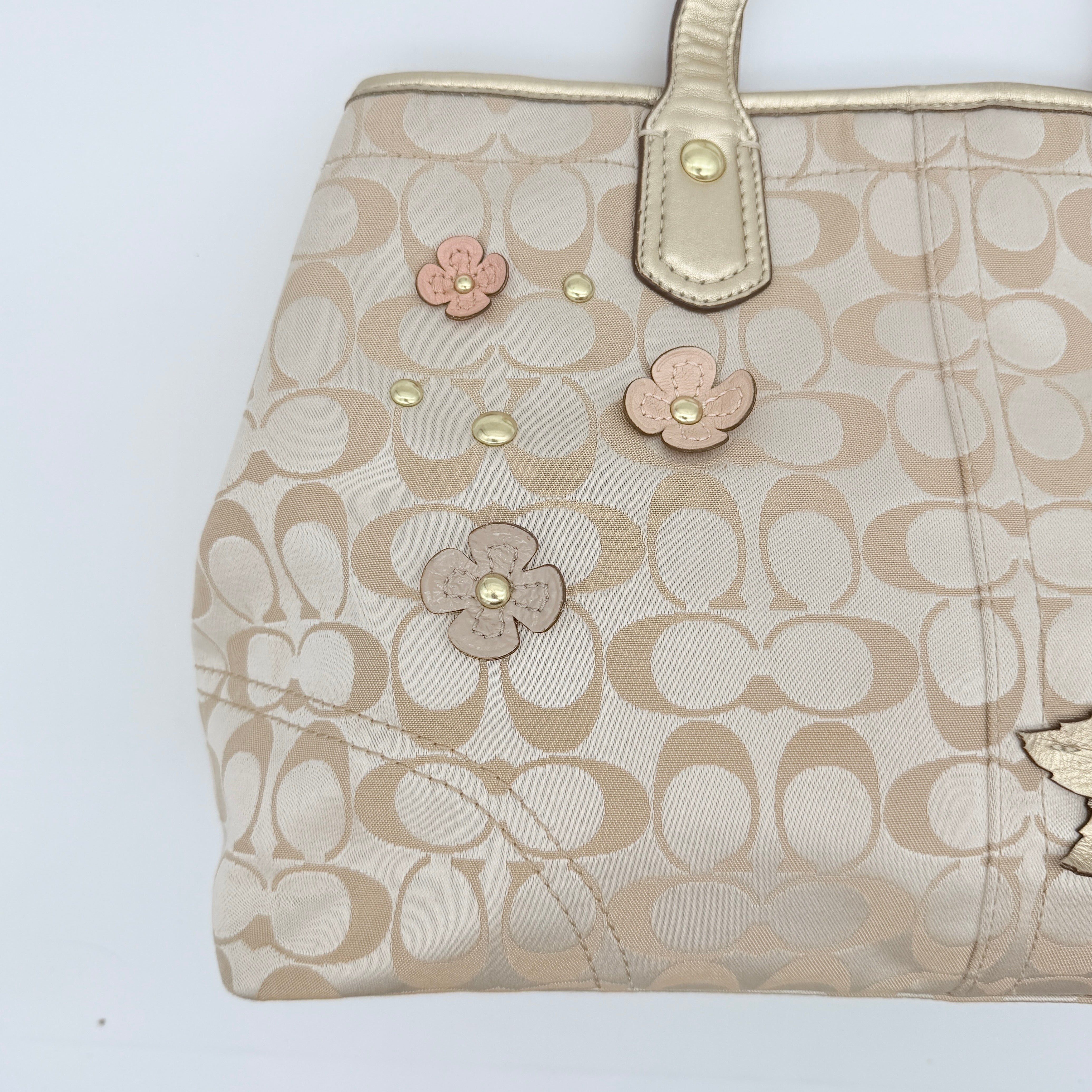 Coach beige carryall tote with floral appliqué and gold-tone handles, styled on a white background.