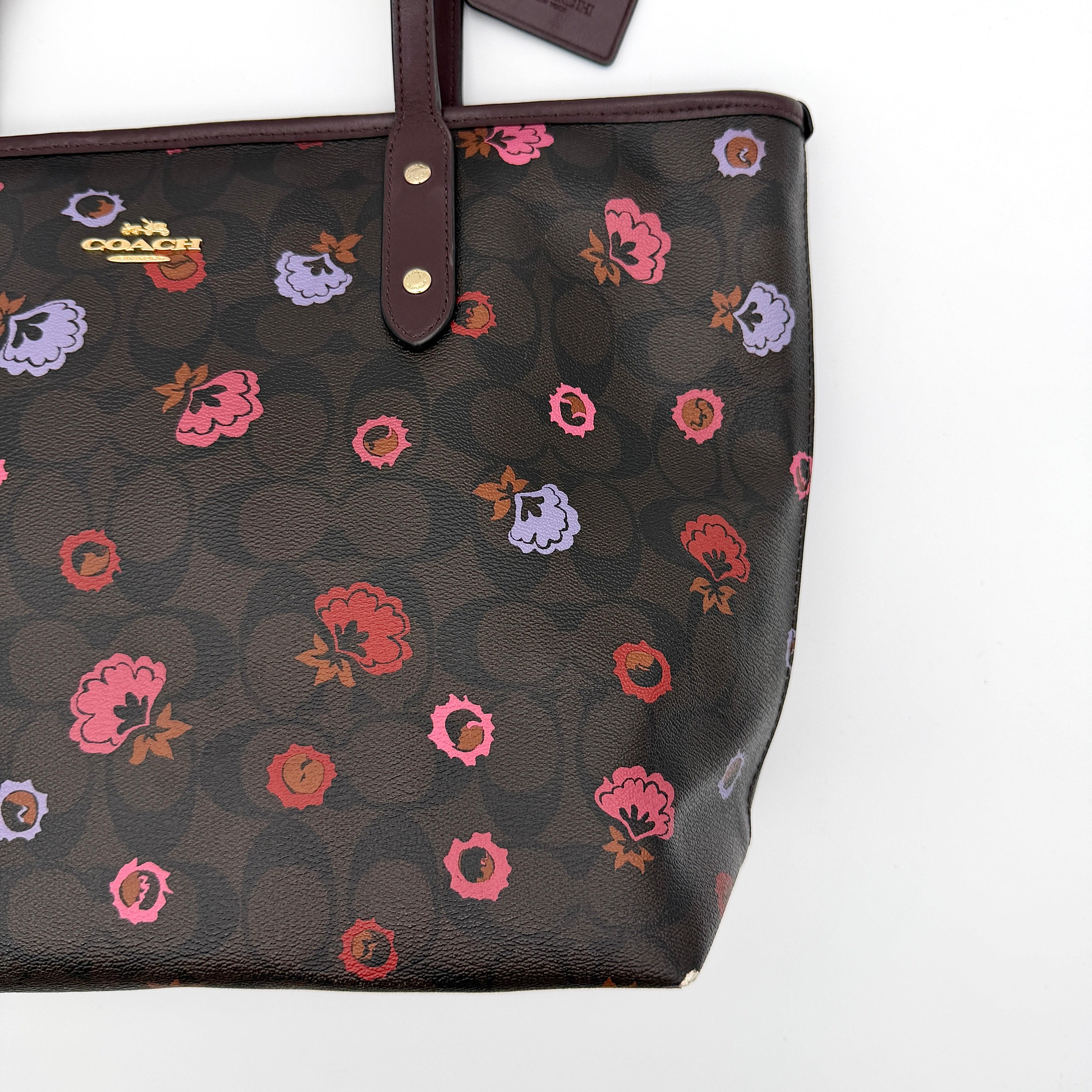 Coach brown coated canvas city tote with pink and lavender floral print, burgundy handles, and gold-tone logo at the front.