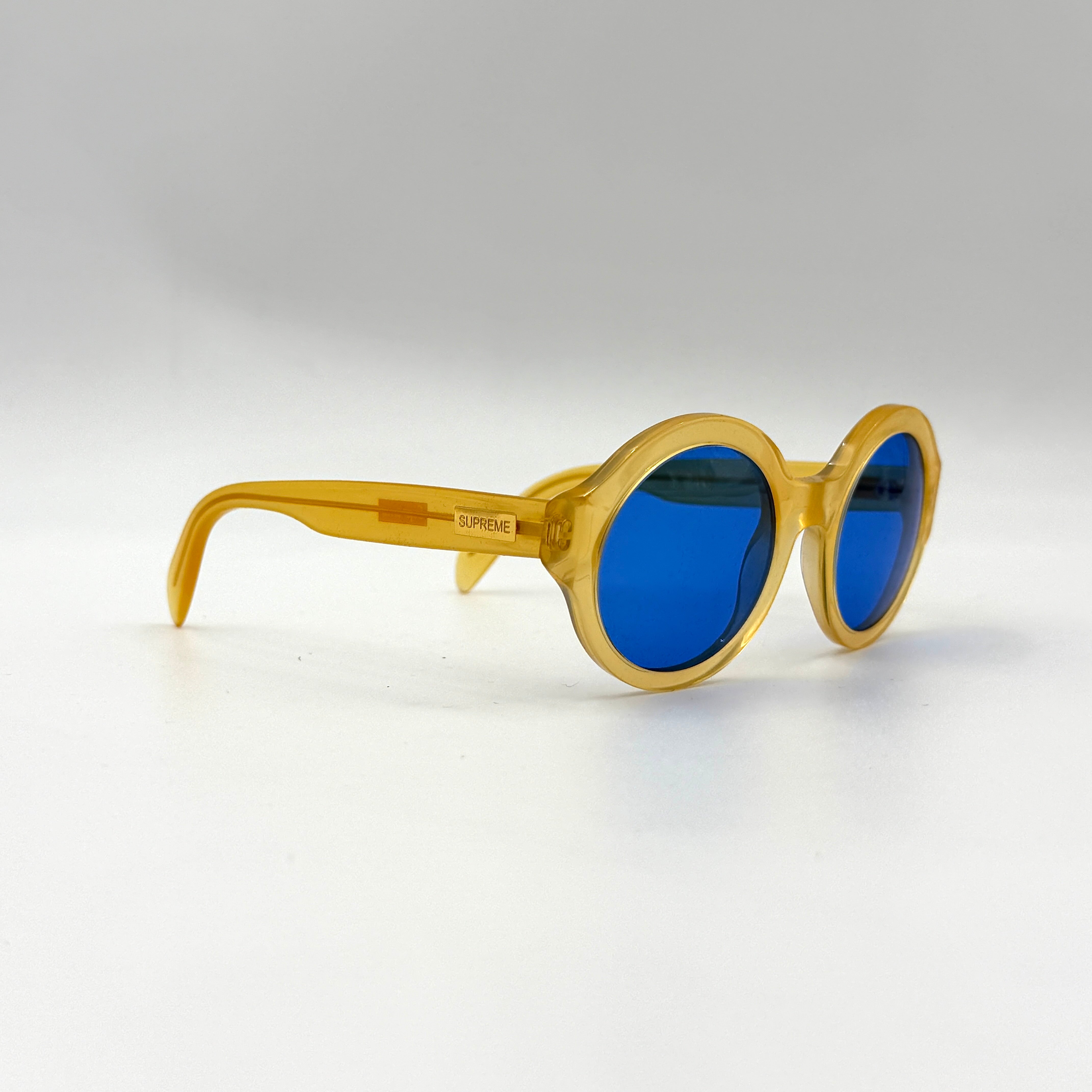 Supreme Downtown Sunglasses SS21 in translucent gold with vivid blue round lenses, set before a black Supreme case.