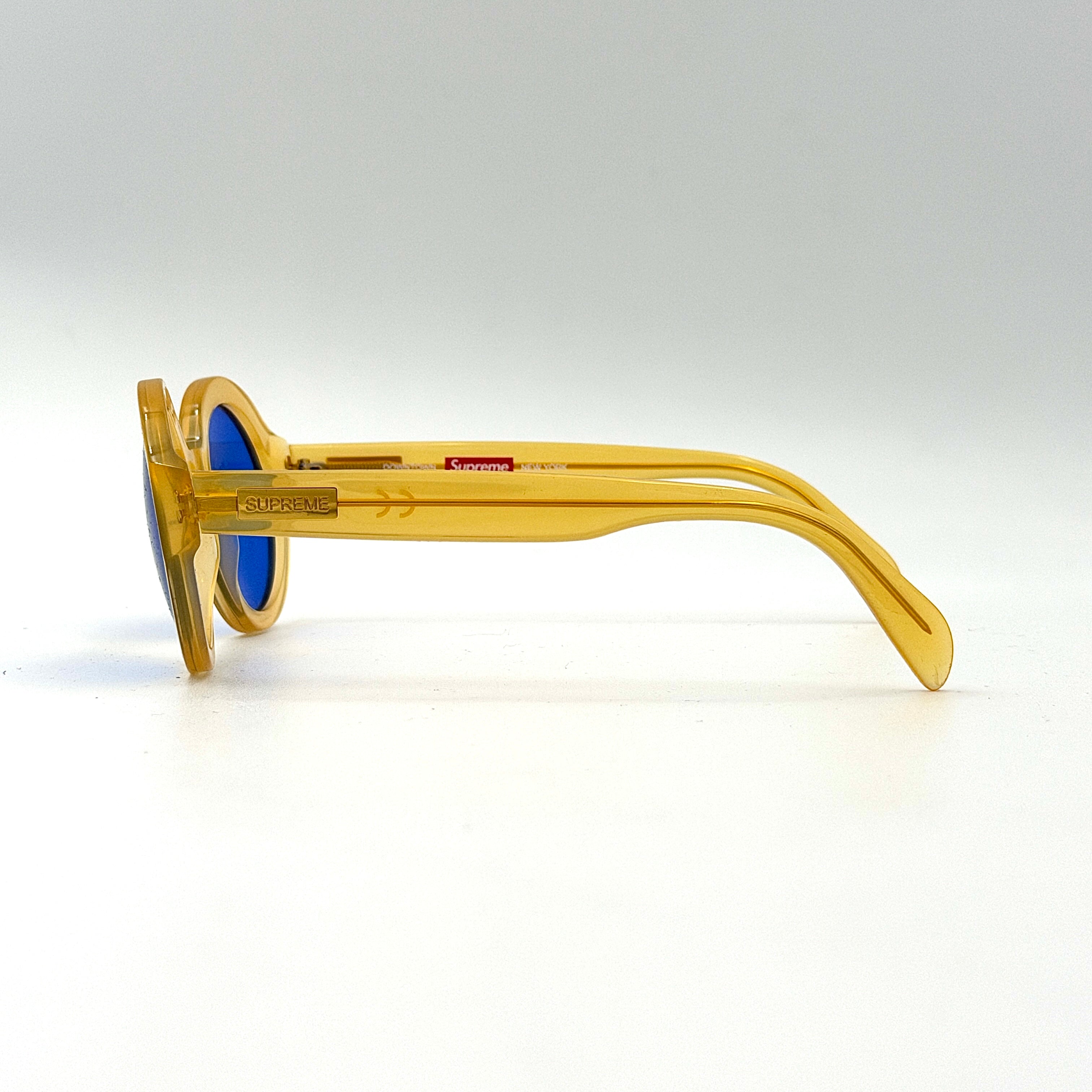 Supreme Downtown Sunglasses SS21 in translucent gold with vivid blue round lenses, set before a black Supreme case.