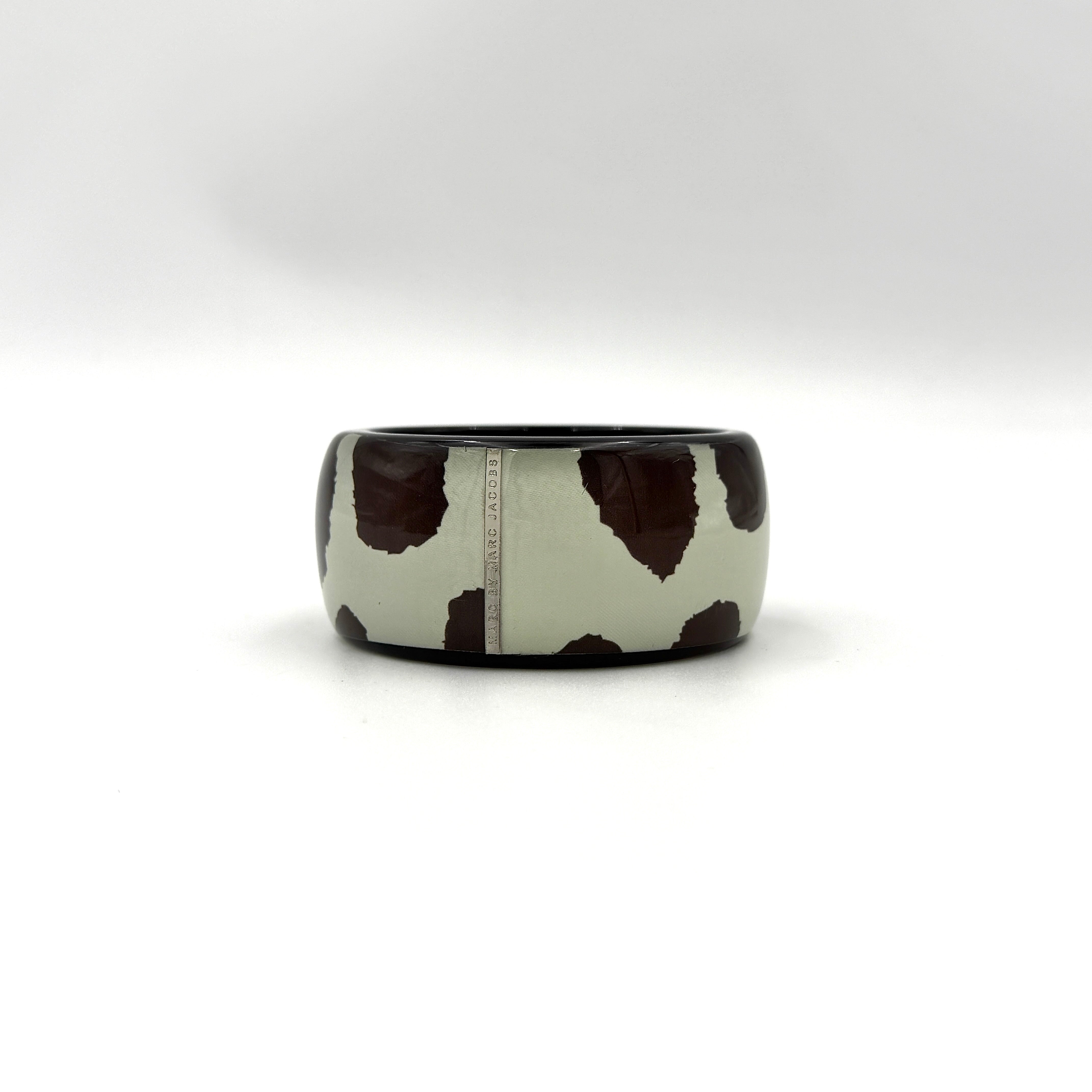 A wide Marc Jacobs bangle bracelet in sage green and dark brown cow print resin, featuring a slim silvertone logo plaque.