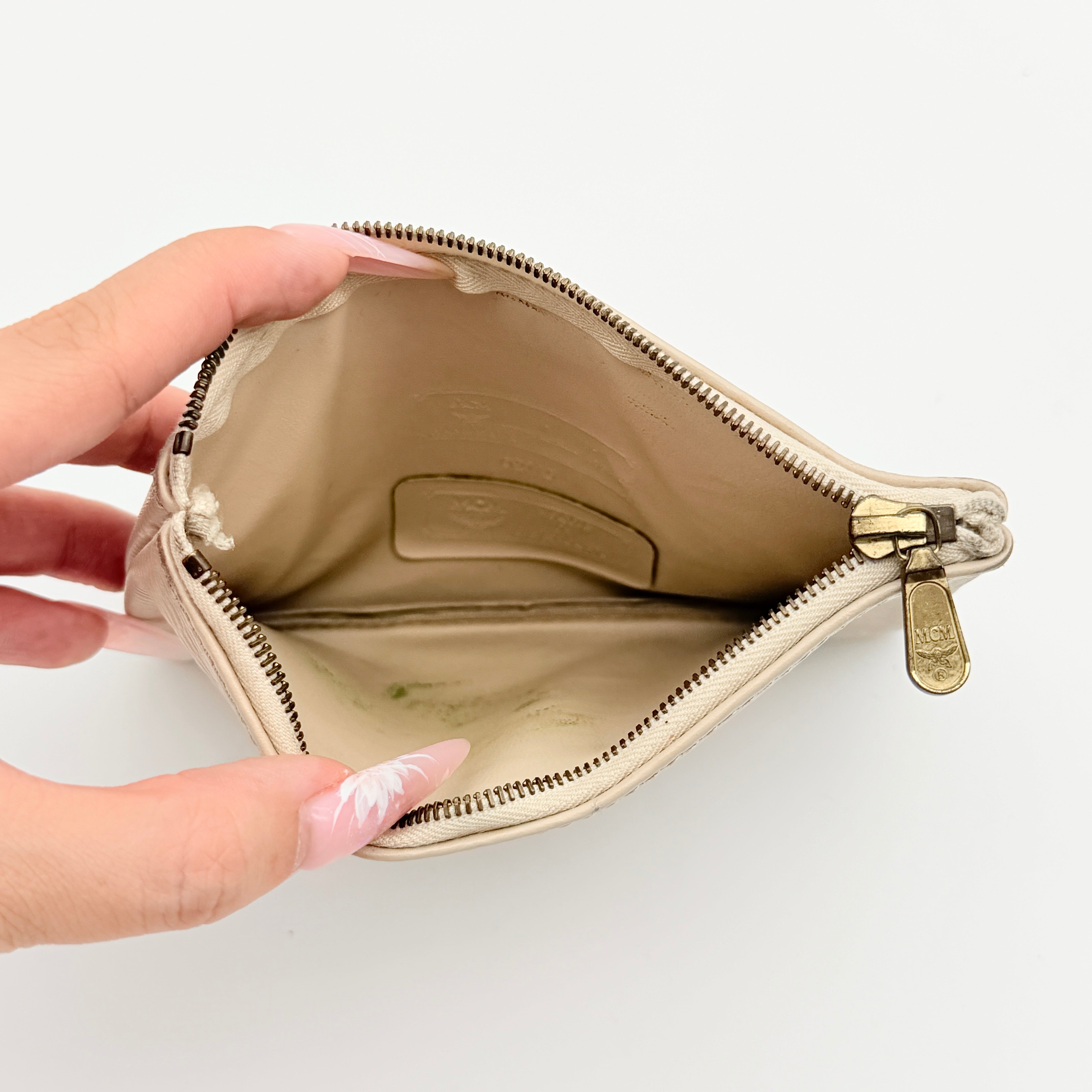 MCM beige embossed leather pouch with iconic gold logo plaque and zip closure, styled against a white background.