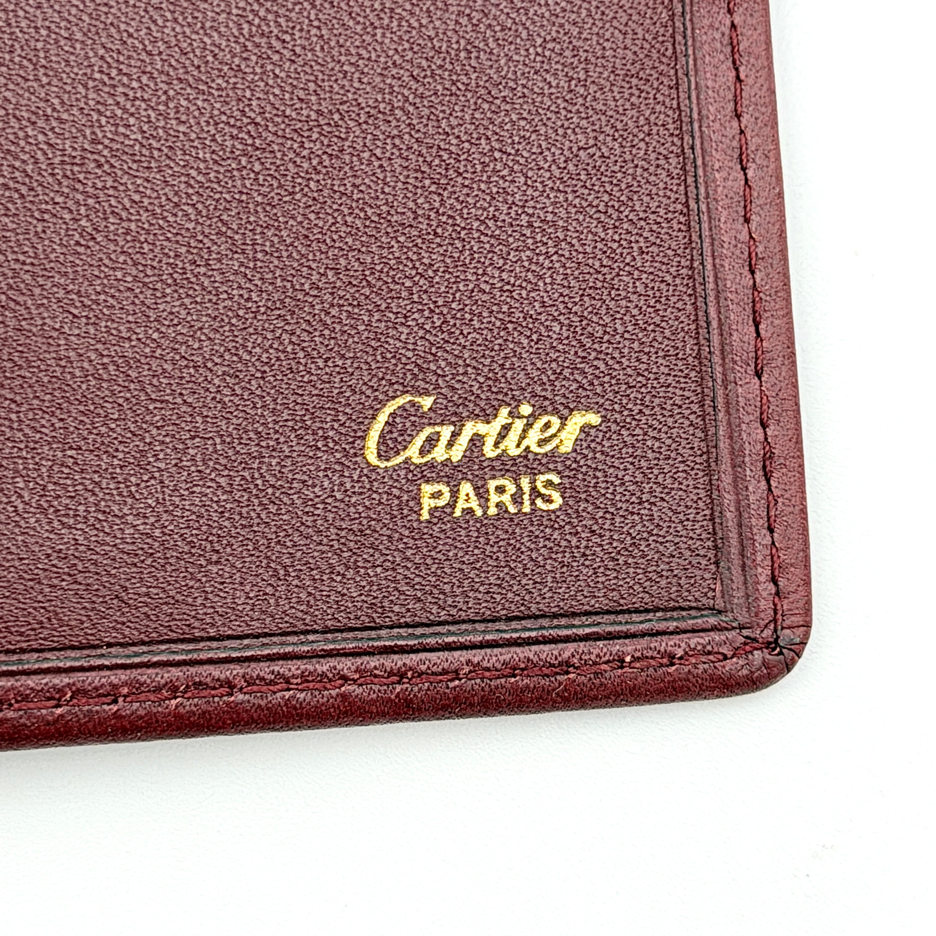 Cartier Must de Cartier leather wallet in rich burgundy with gold-tone corner accents and an embossed logo, photographed closed against a white background.