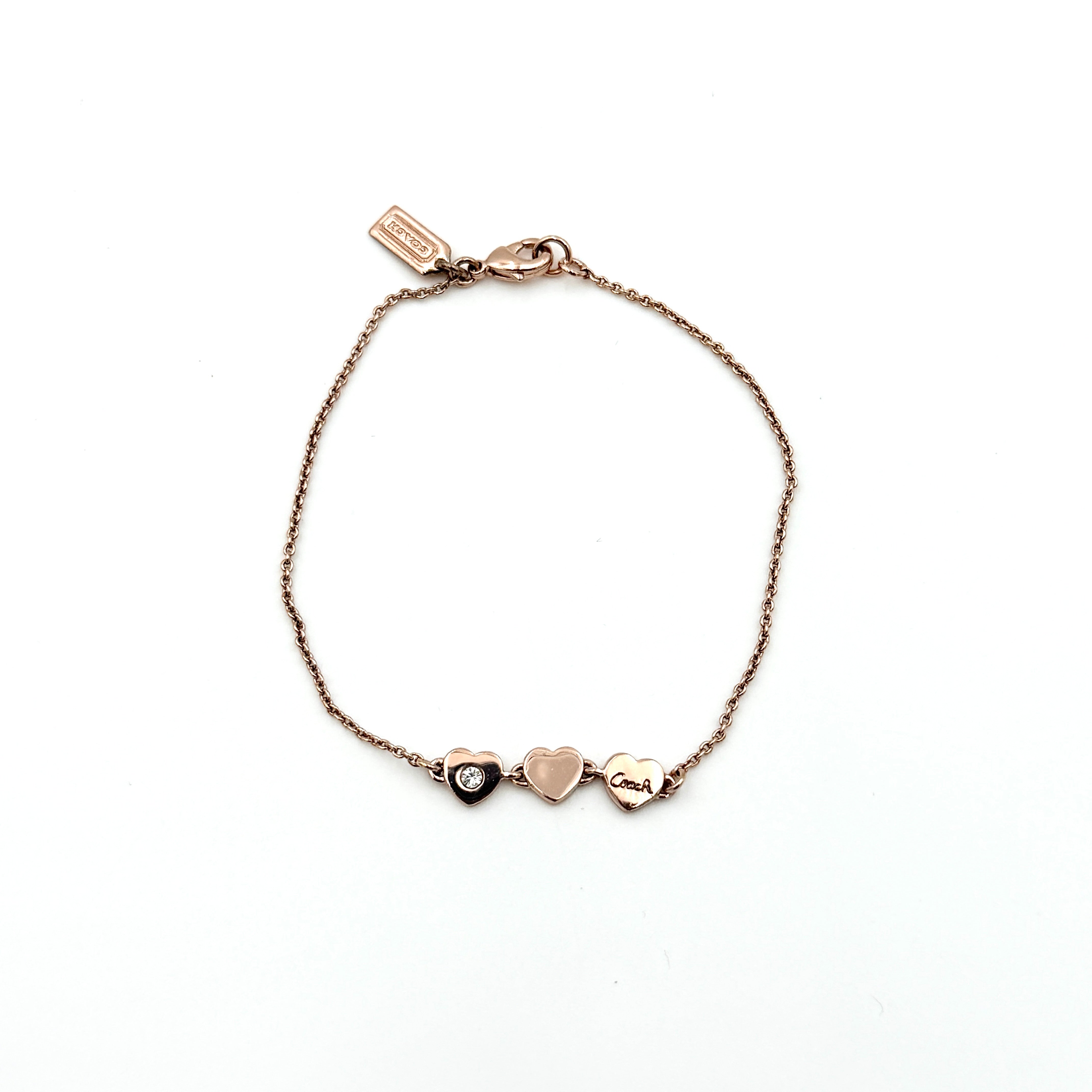 Coach gold bracelet featuring three heart-shaped charms, one with a crystal, one plain, and one engraved with the Coach logo.