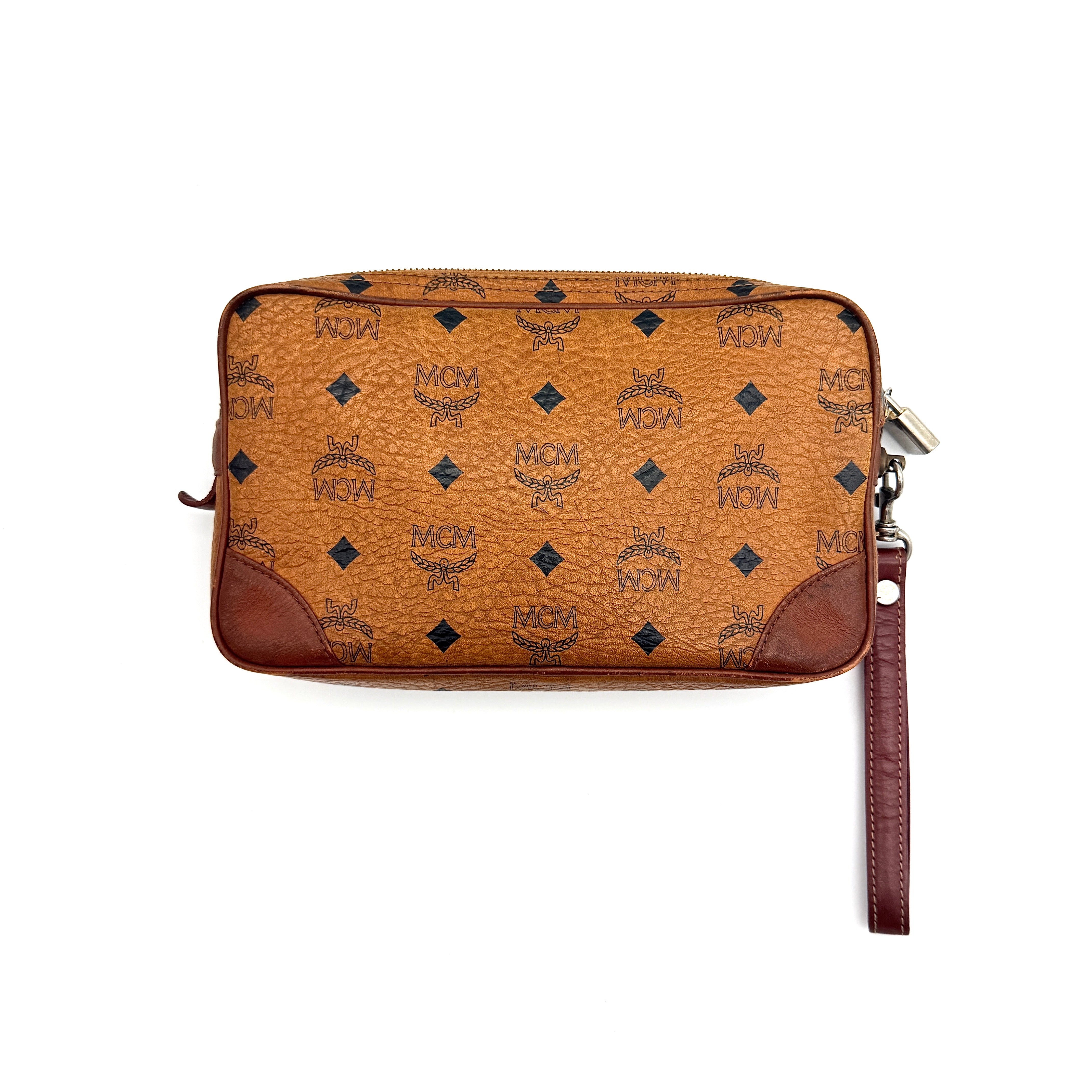 MCM Visetos monogram clutch bag in brown coated canvas with wristlet strap and gold logo plaque, photographed on white background.