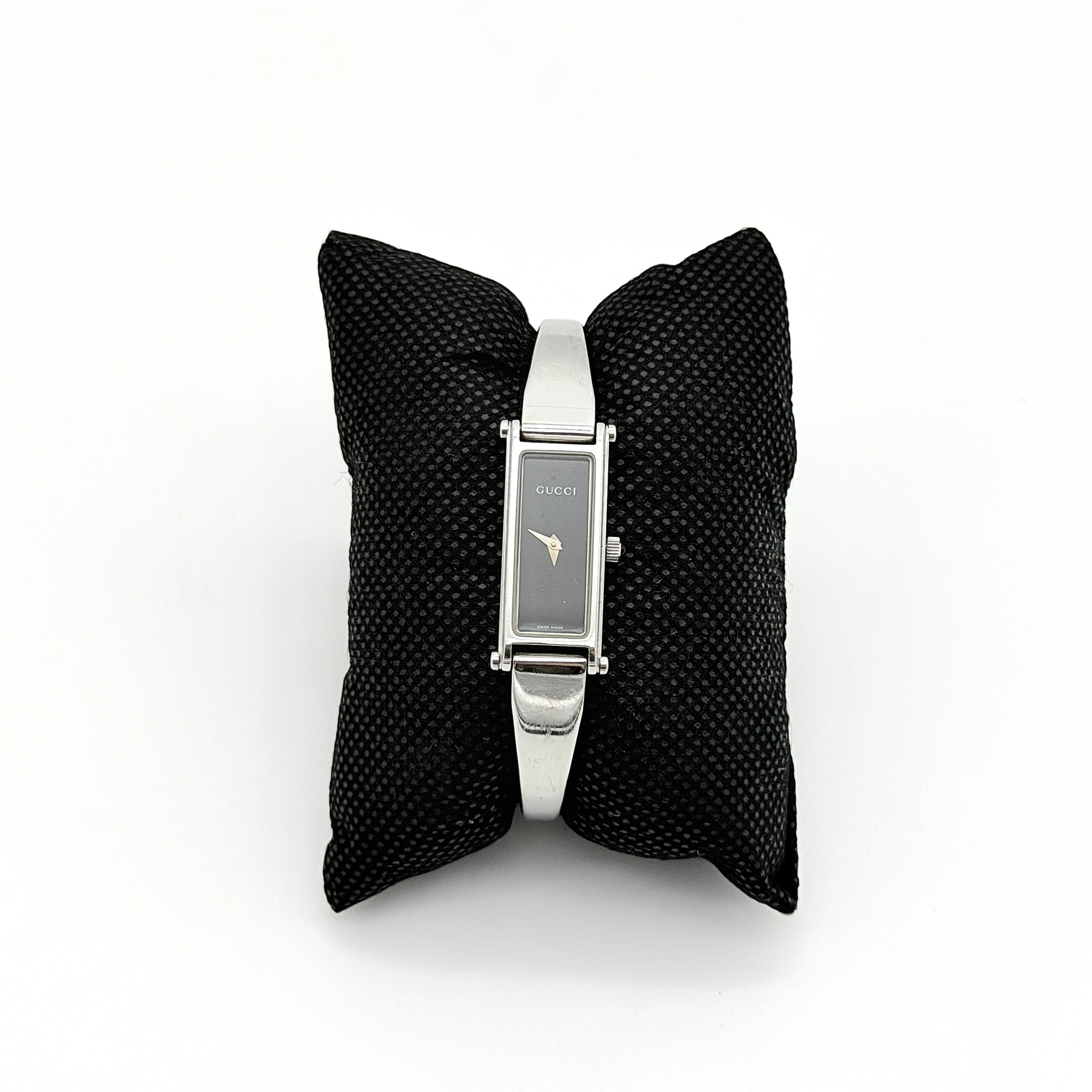 Gucci 1500L black dial bangle watch with stainless steel bracelet, photographed on a clear display stand.