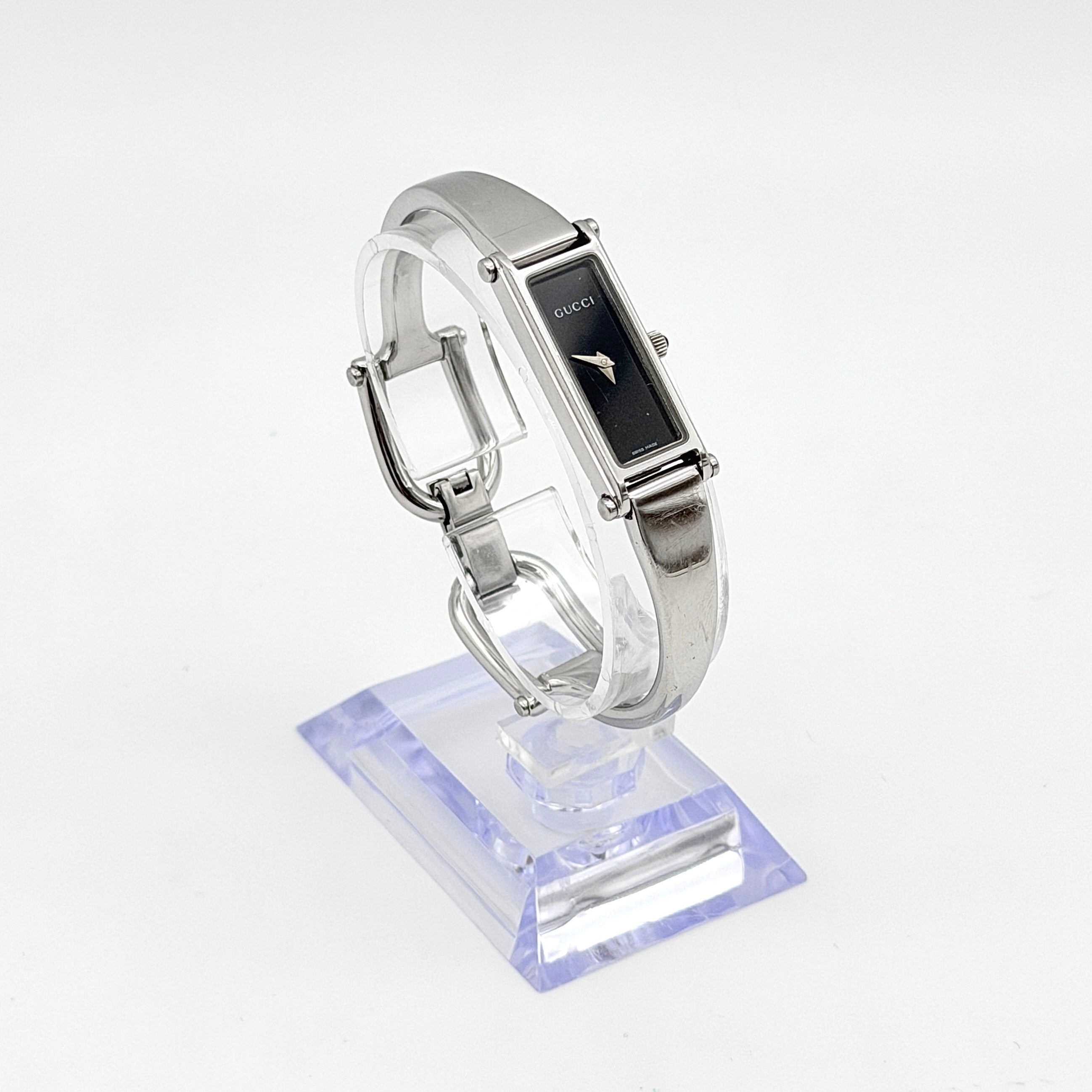 Gucci 1500L black dial bangle watch with stainless steel bracelet, photographed on a clear display stand.