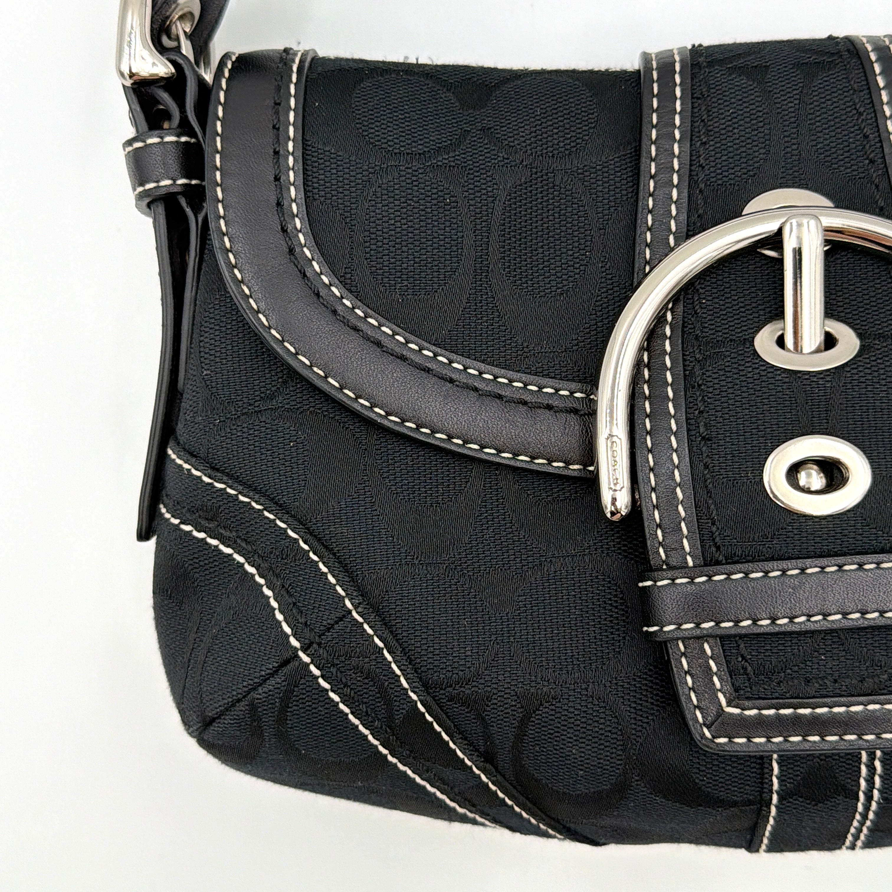 Coach black Signature Soho shoulder bag with oversized silver buckle, cream contrast stitching, and monogram jacquard canvas backdrop.