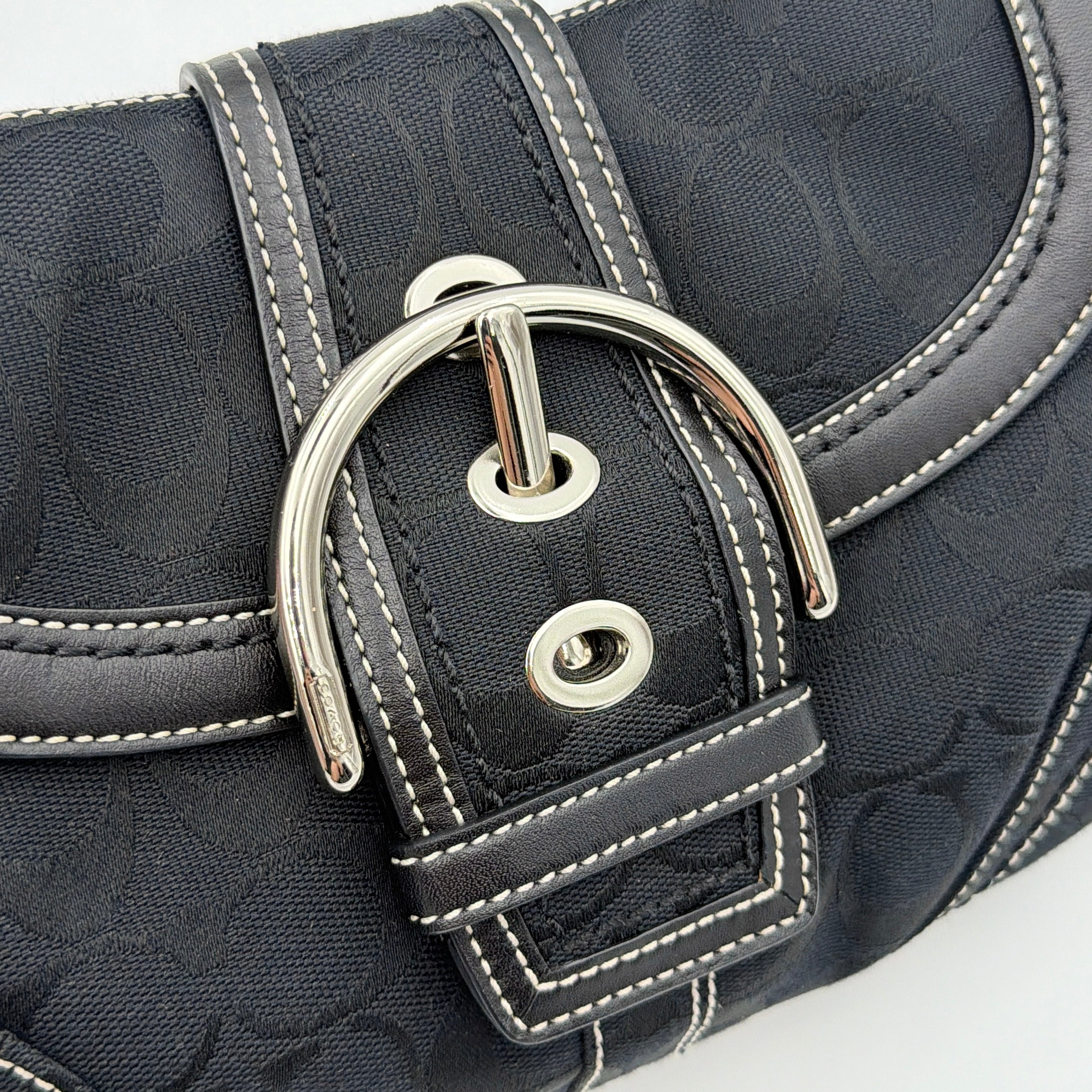 Coach black Signature Soho shoulder bag with oversized silver buckle, cream contrast stitching, and monogram jacquard canvas backdrop.