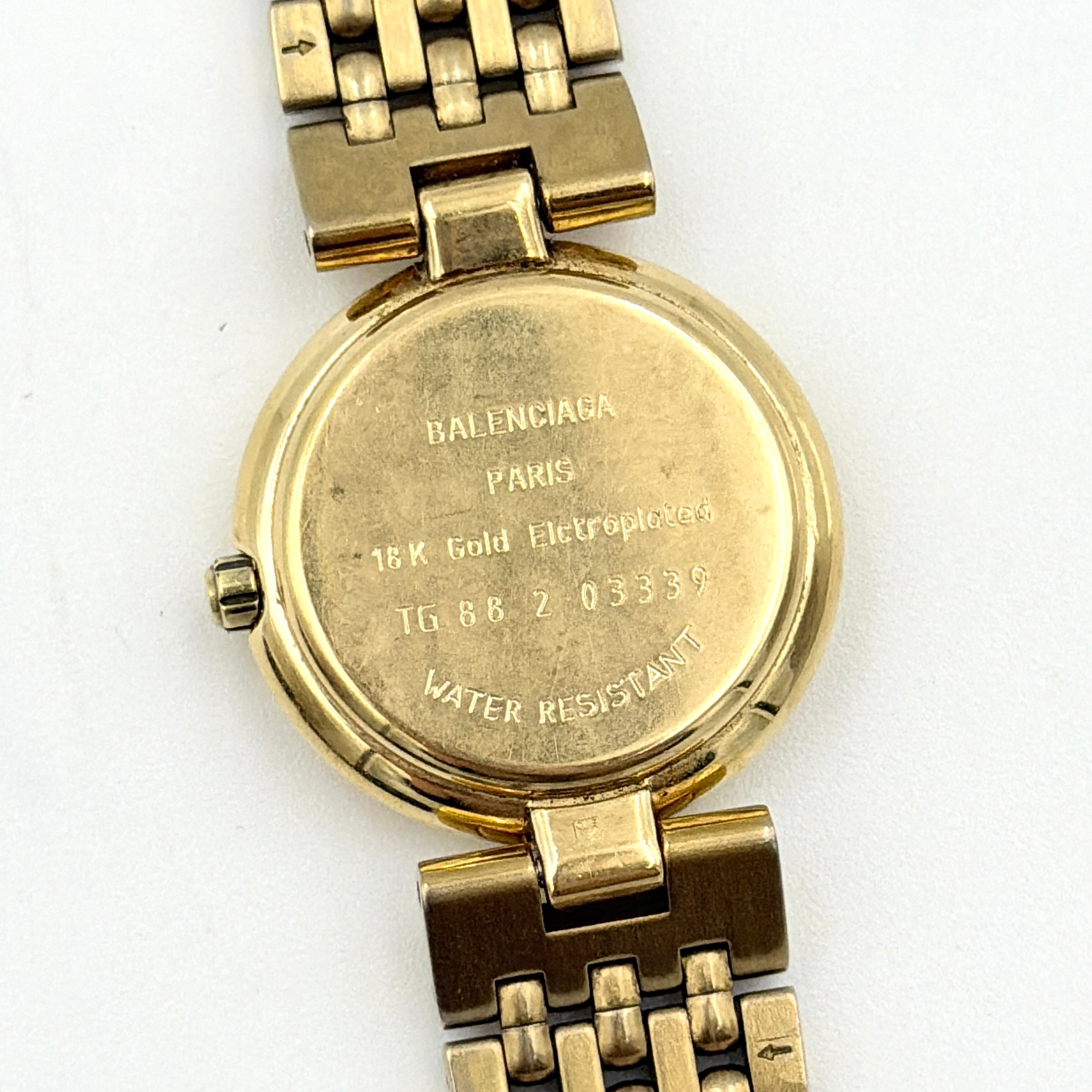 Balenciaga BB Quartz gold watch with a round dial, diamond-shaped hour markers, and a gold link bracelet displayed on a black cushion.