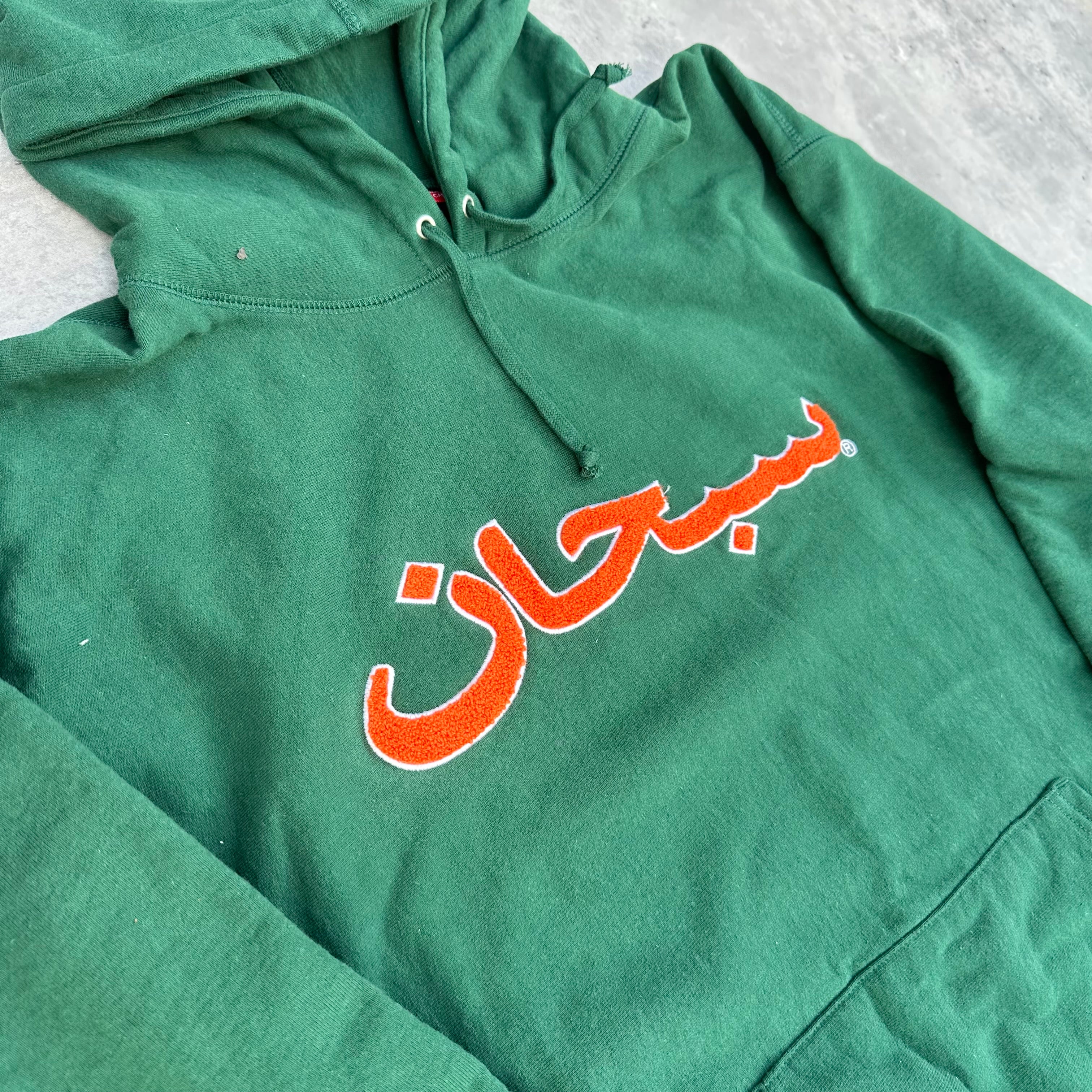 Supreme Arabic Logo Hoodie FW21 in green, featuring orange Arabic logo embroidery and kangaroo pocket, made from heavyweight cotton.
