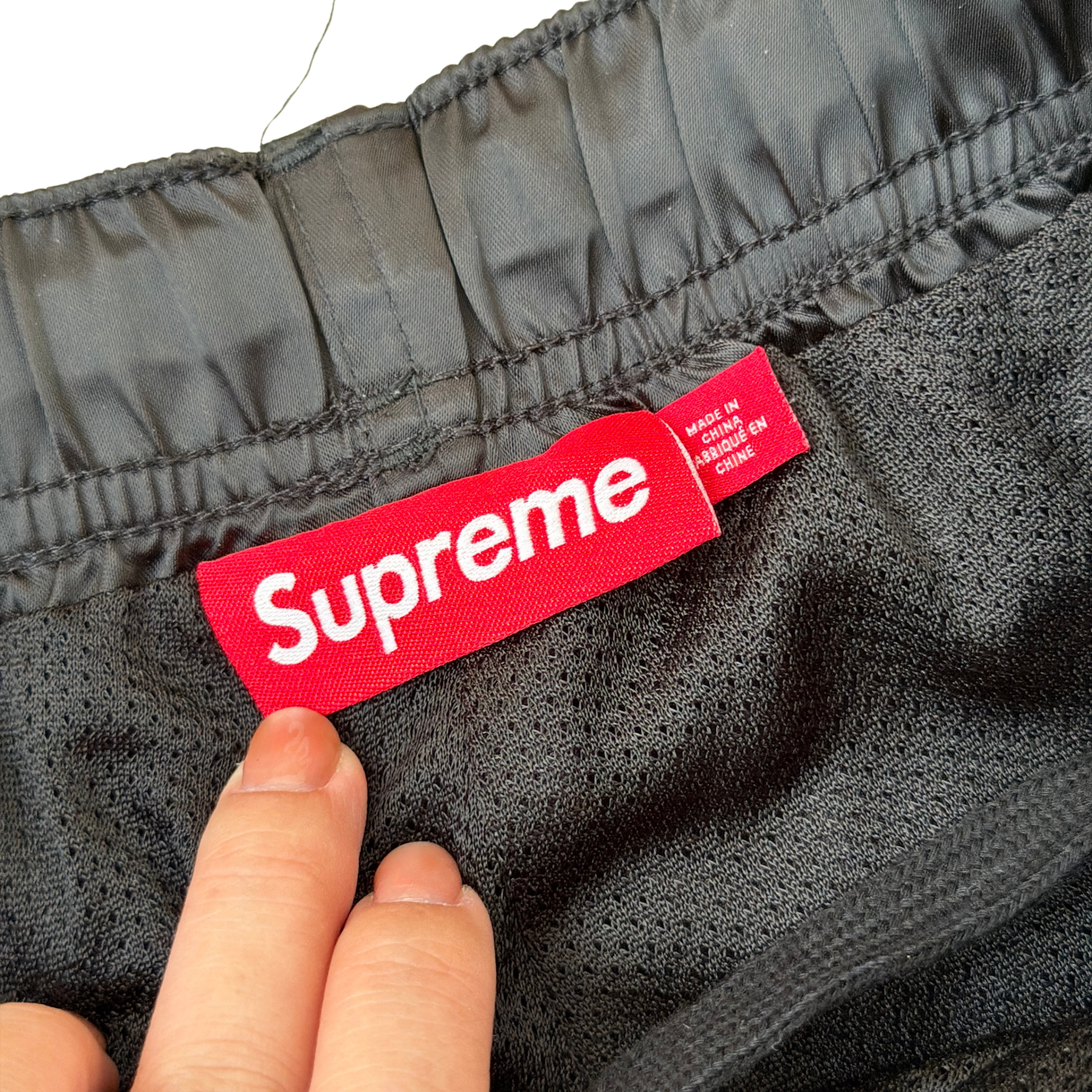 Supreme black satin track pants with white piping, a crest and Supreme logo embroidery at the hip, styled with relaxed legs.