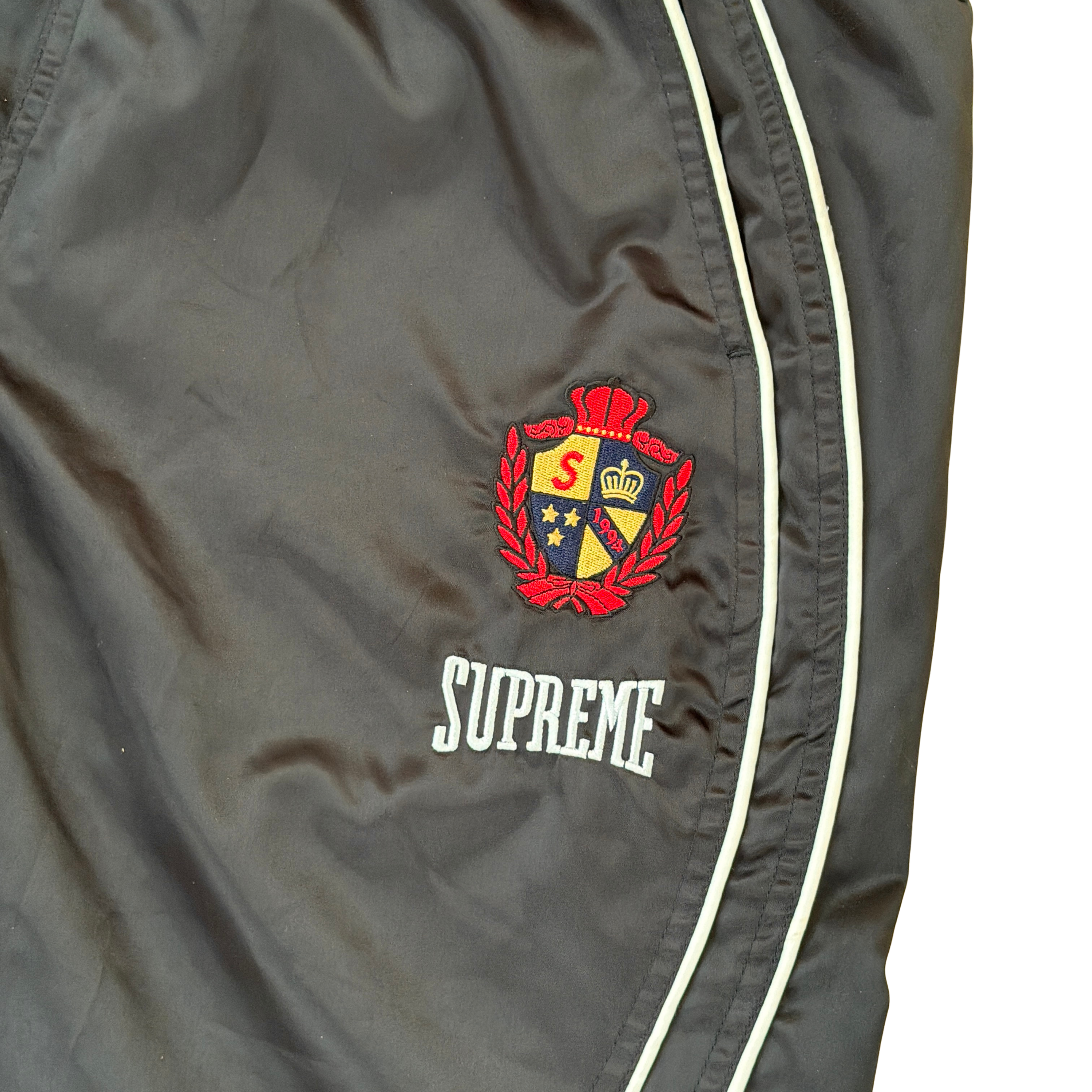 Supreme black satin track pants with white piping, a crest and Supreme logo embroidery at the hip, styled with relaxed legs.