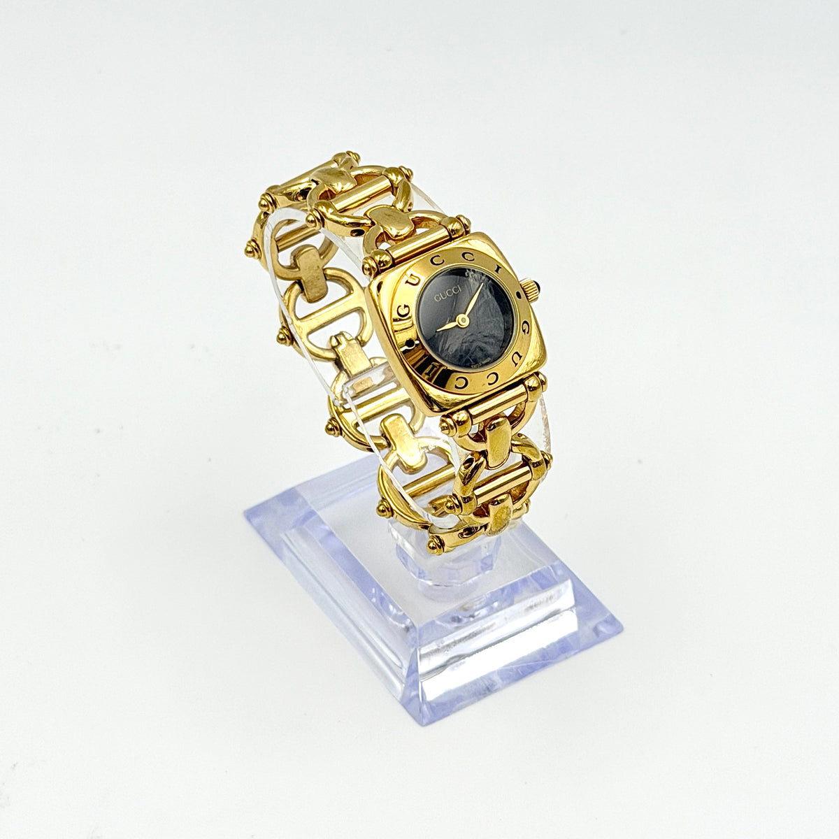 Gucci 6400L gold-tone bracelet watch with a black dial and distinctive horsebit links, showcased on a clear stand.