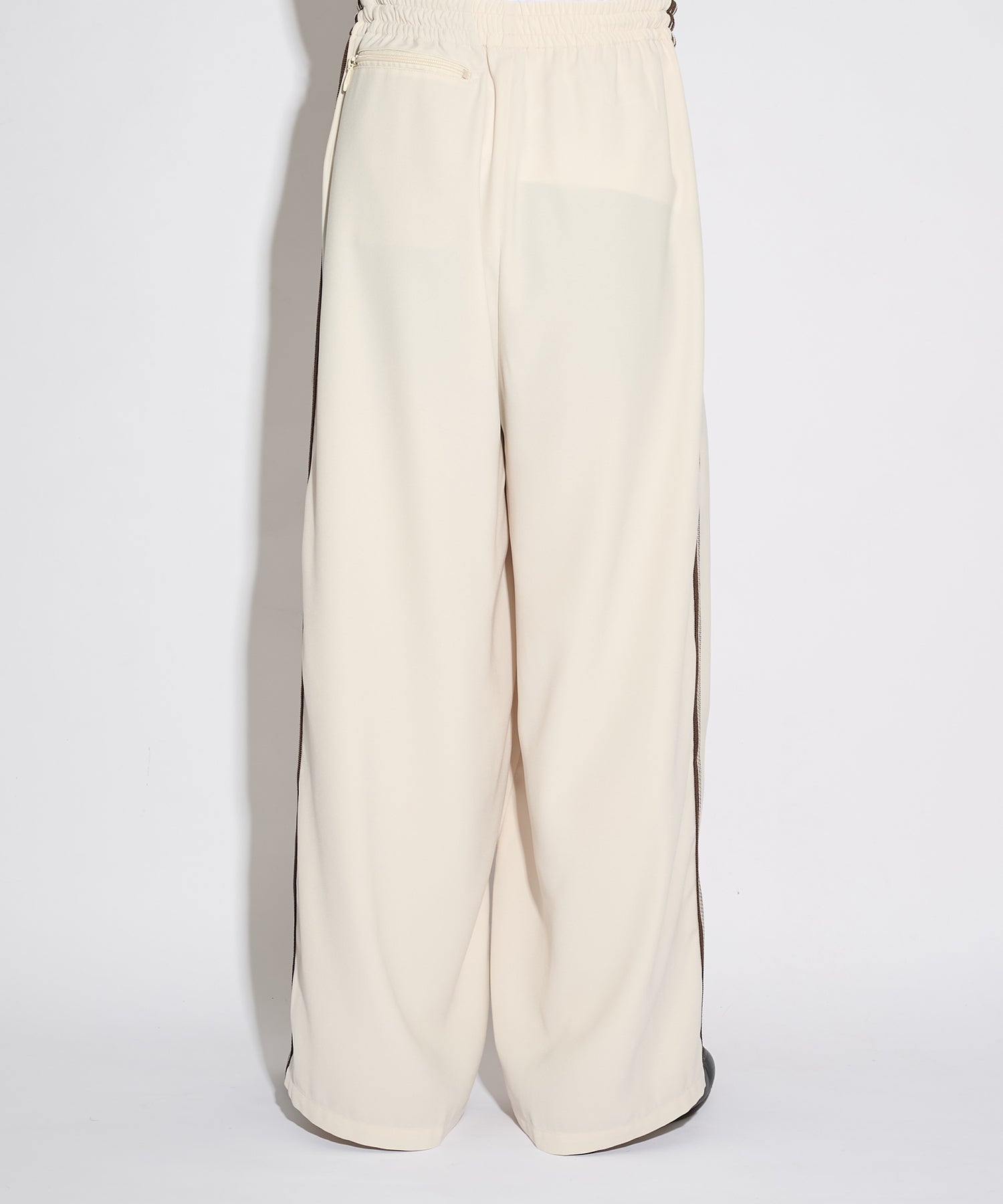 Needles H.D. Track Pant in cream with brown side stripes and embroidered butterfly logo, shown from the side in soft studio lighting.