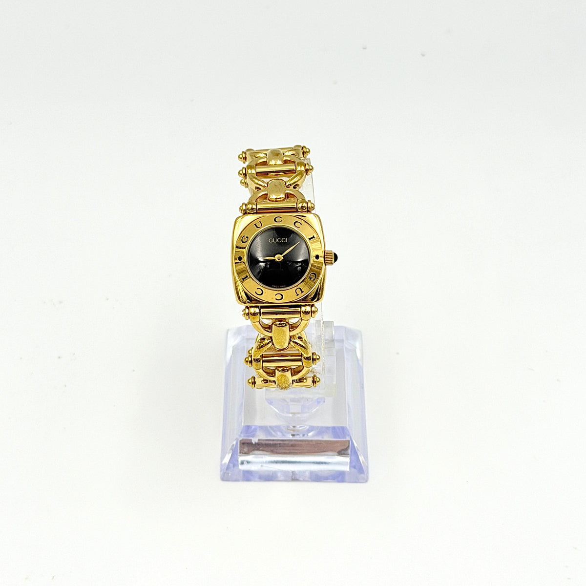 Gucci 6400L gold-tone bracelet watch with a black dial and distinctive horsebit links, showcased on a clear stand.