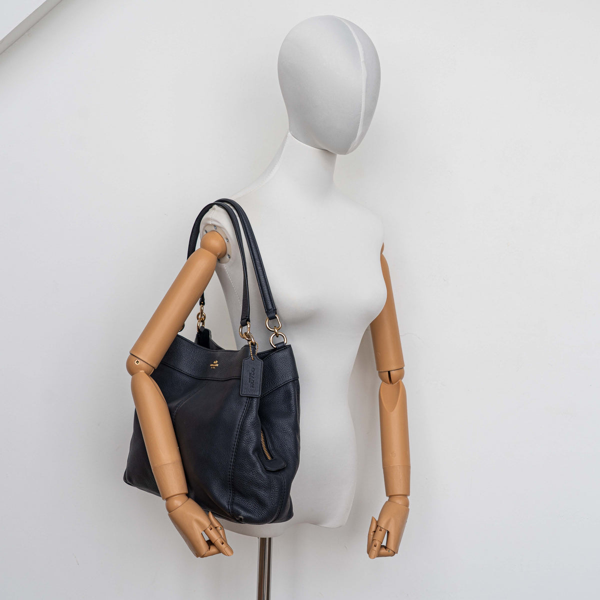 Coach Lexy Shoulder Bag in black pebbled leather with gold-tone hardware and double handles, styled against a white background.