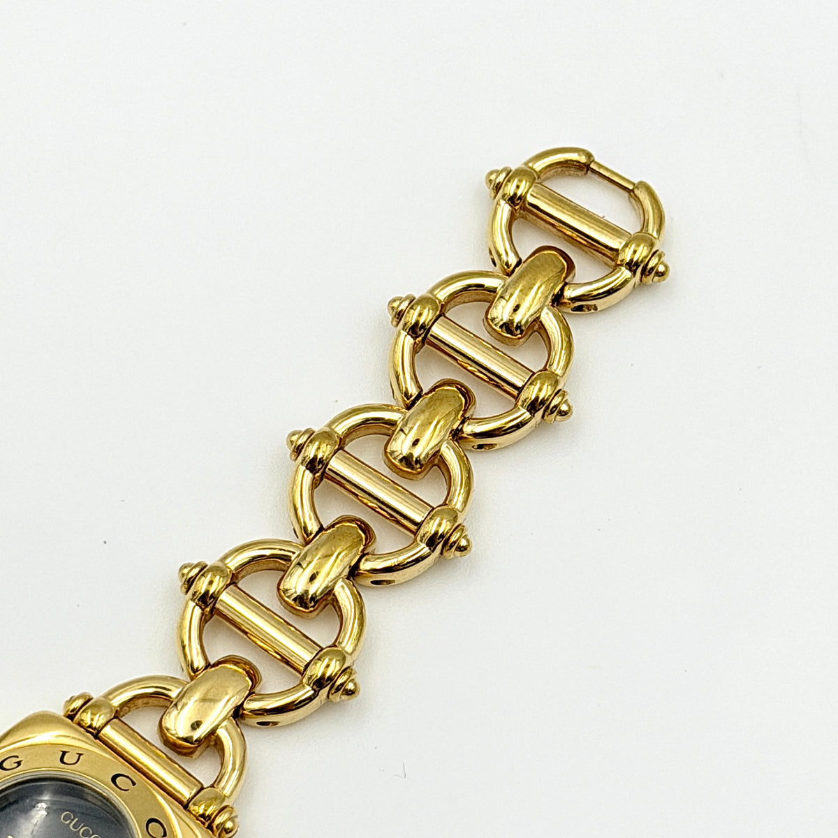 Gucci 6400L gold-tone bracelet watch with a black dial and distinctive horsebit links, showcased on a clear stand.