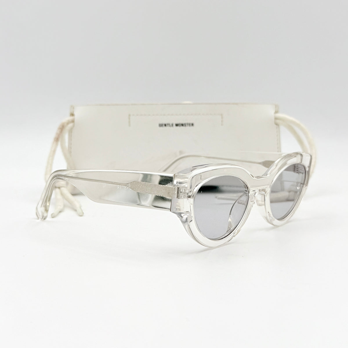 Gentle Monster Tazi transparent sunglasses with grey lenses and clear acetate frame, softly styled alongside the branded case.