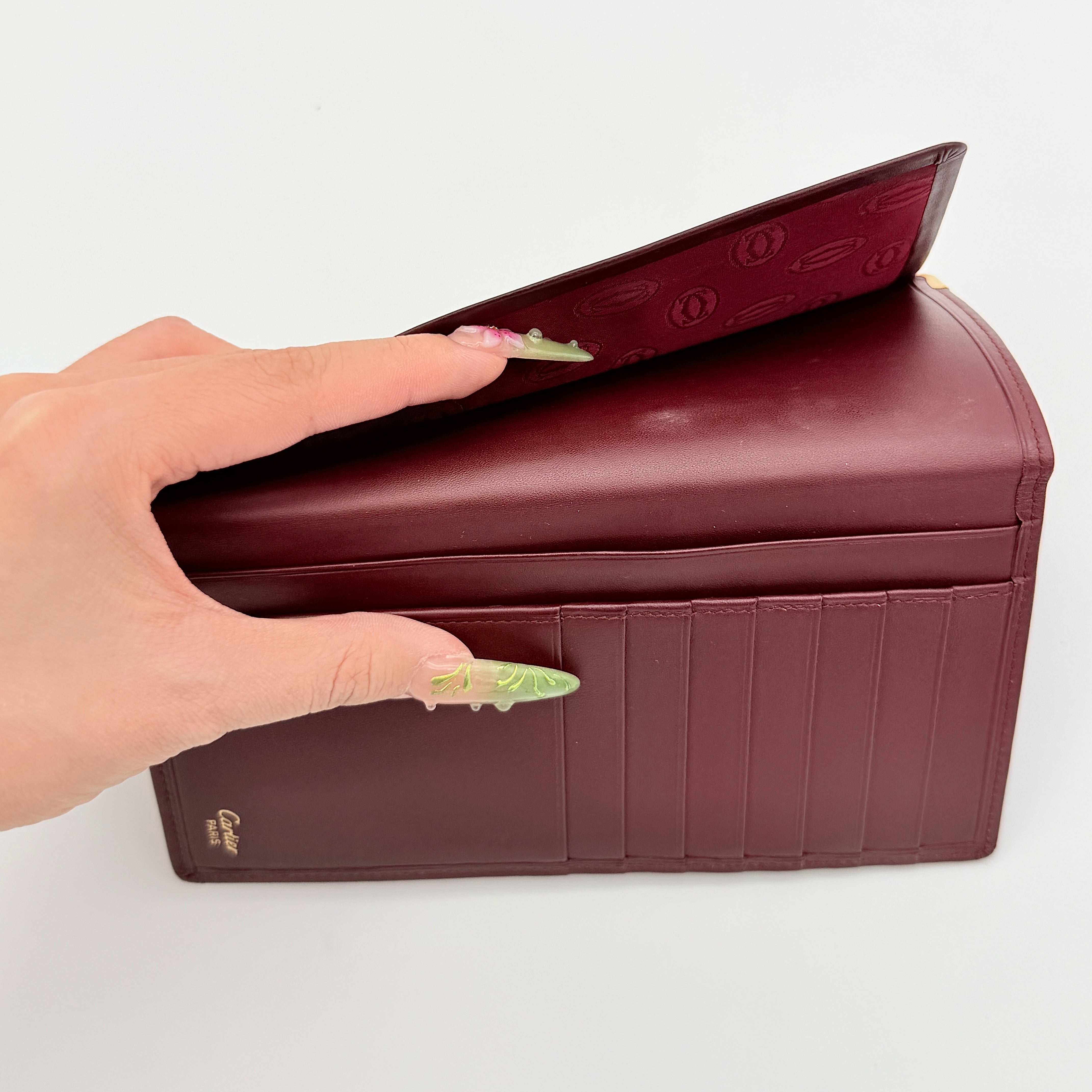 Cartier Must de Cartier bifold long wallet in burgundy leather with gold-tone corner accents, shown closed against a white background.