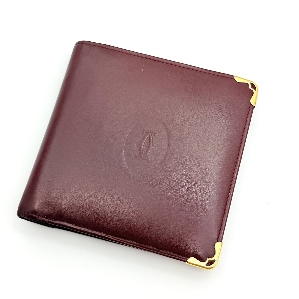 Cartier Must de Cartier bifold wallet in burgundy leather with gold-tone corners and embossed logo, photographed against a white background.