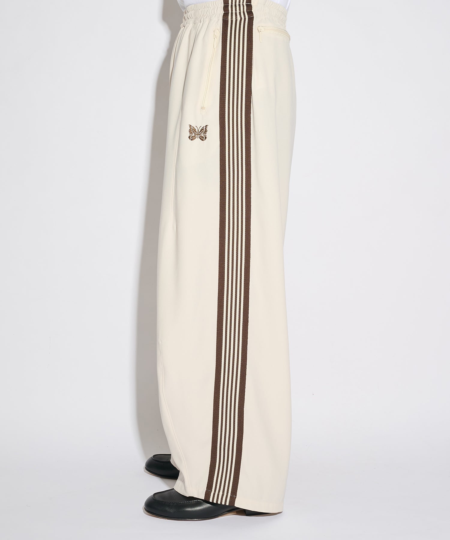 Needles H.D. Track Pant in cream with brown side stripes and embroidered butterfly logo, shown from the side in soft studio lighting.
