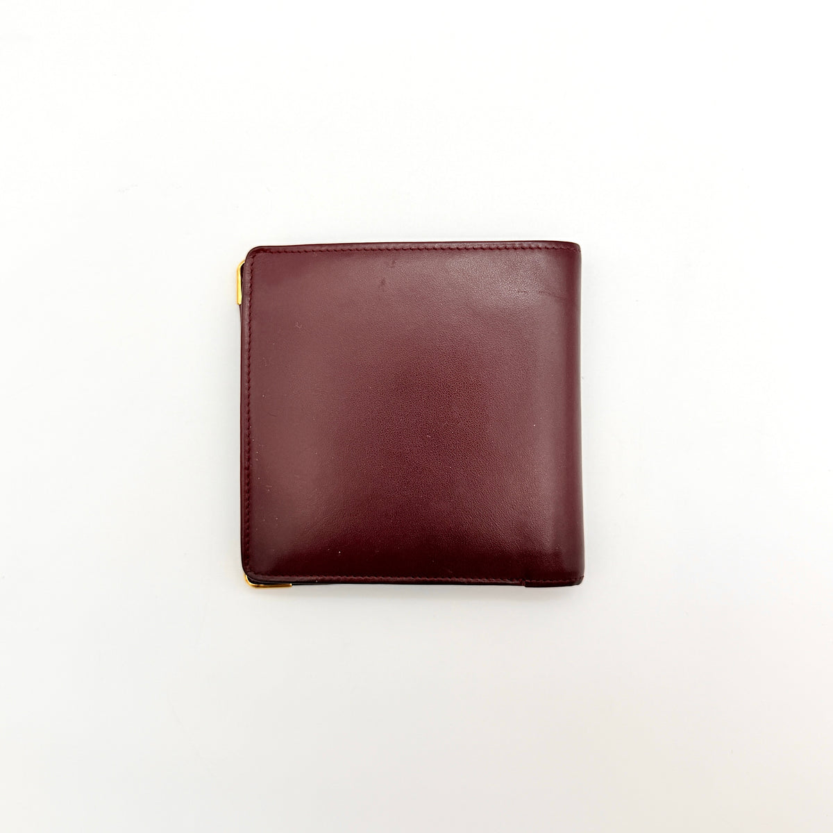 Cartier Must de Cartier bifold wallet in burgundy leather with gold-tone corners and embossed logo, photographed against a white background.