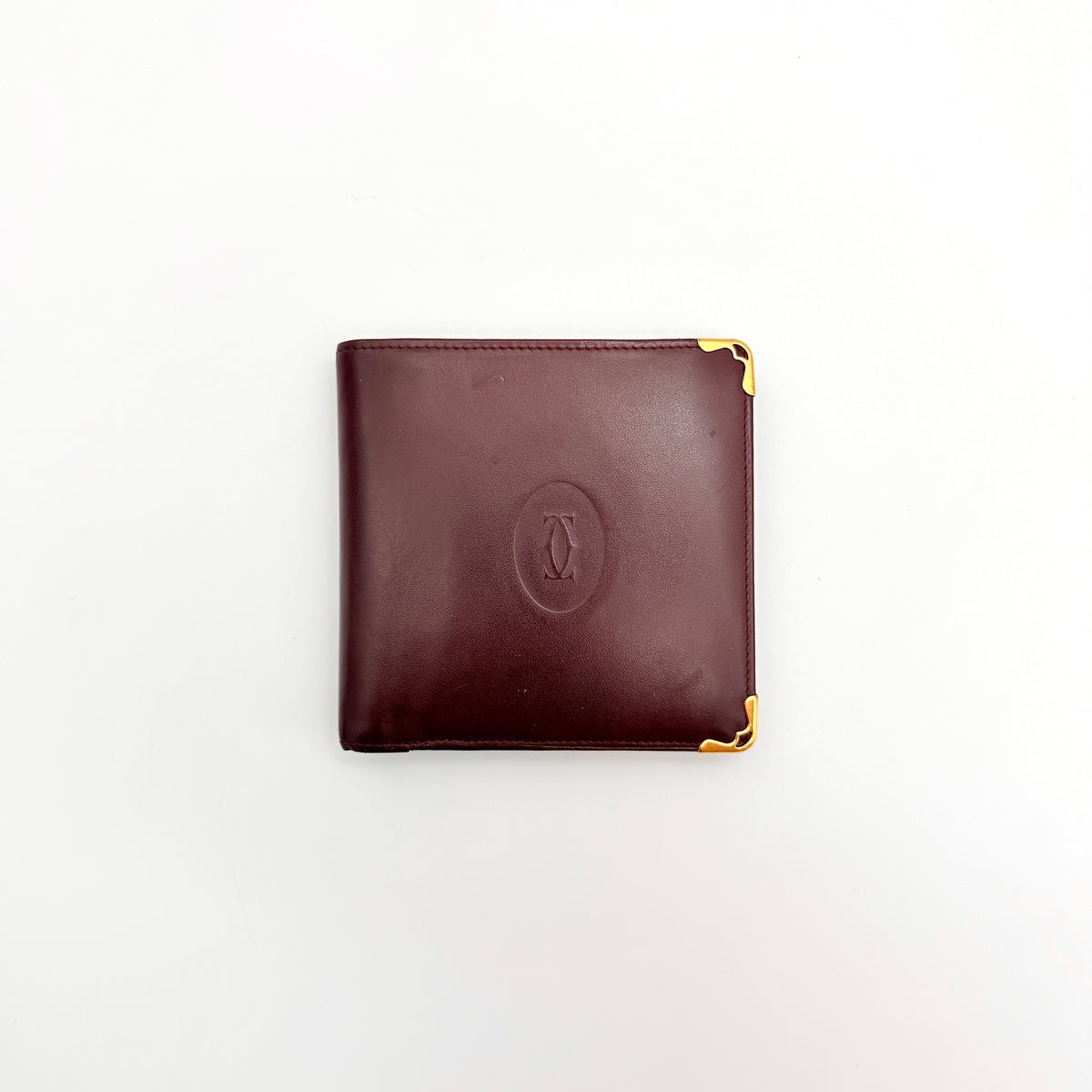 Cartier Must de Cartier bifold wallet in burgundy leather with gold-tone corners and embossed logo, photographed against a white background.