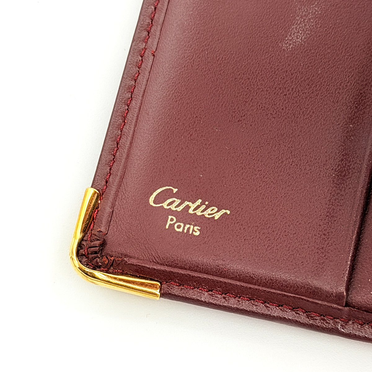 Cartier Must de Cartier bifold wallet in burgundy leather with gold-tone corners and embossed logo, photographed against a white background.