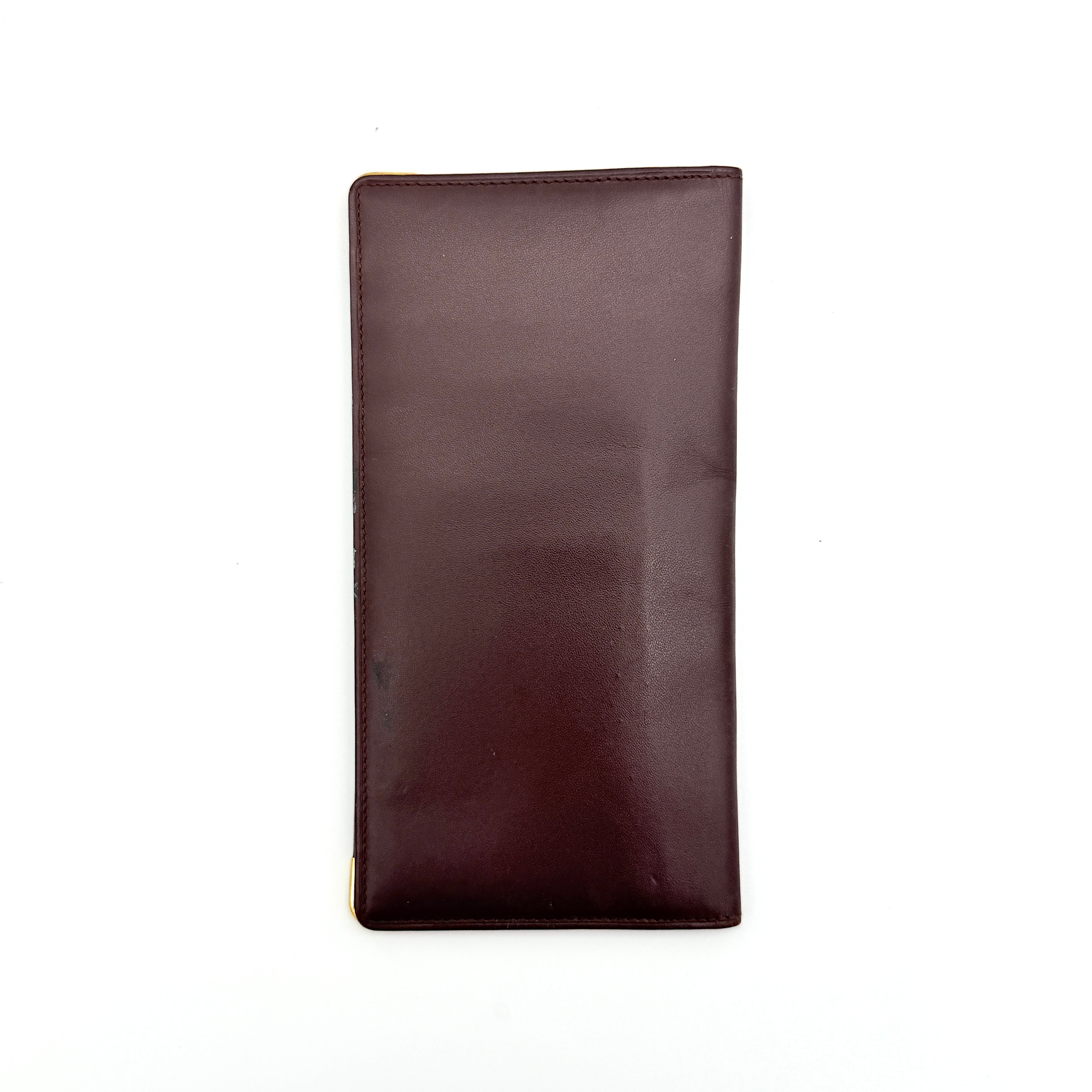Cartier Must de Cartier bifold long wallet in burgundy leather with gold-tone corner accents, shown closed against a white background.