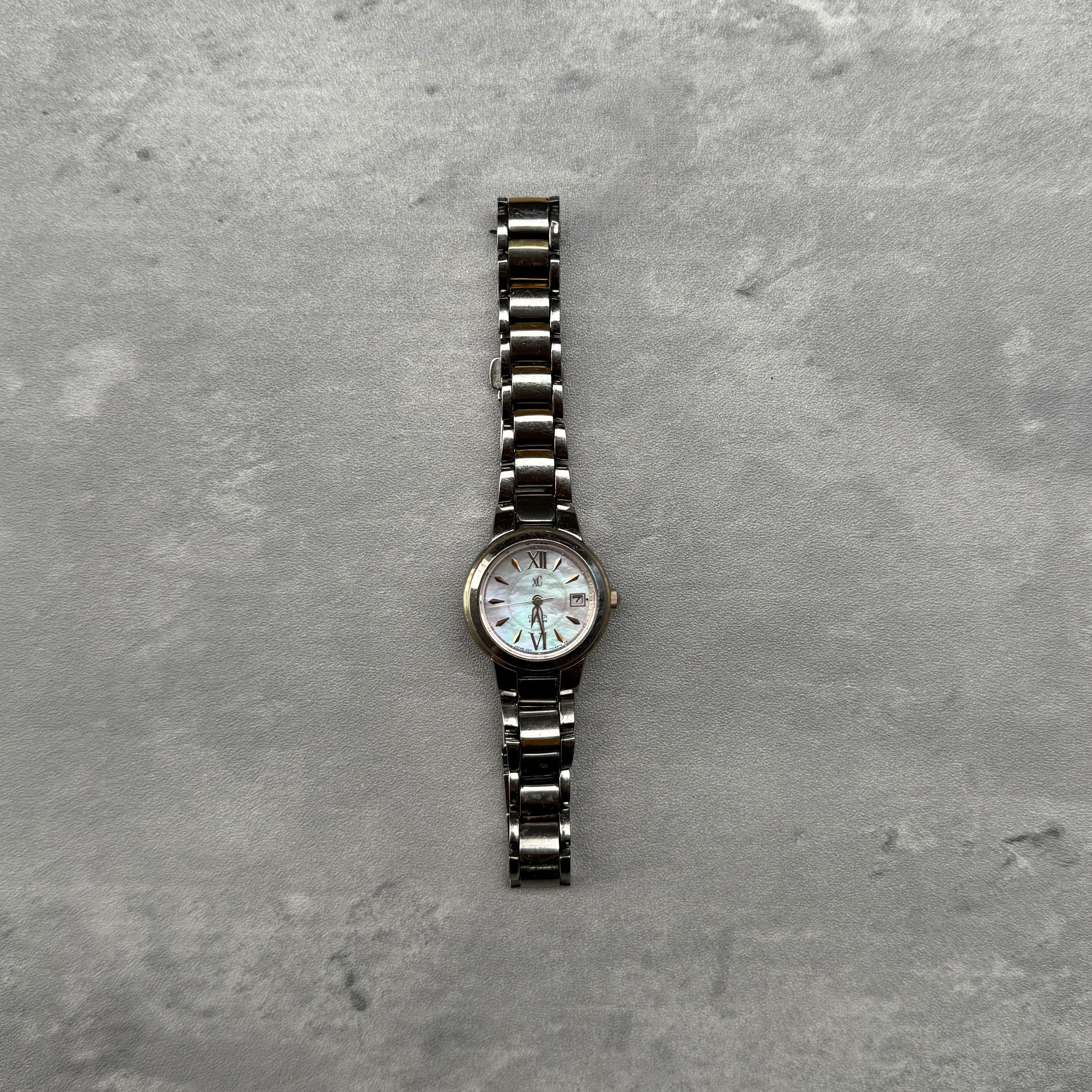 Citizen XC stainless steel watch with mother-of-pearl dial, Roman numeral markers, gold-tone indices, and date display.