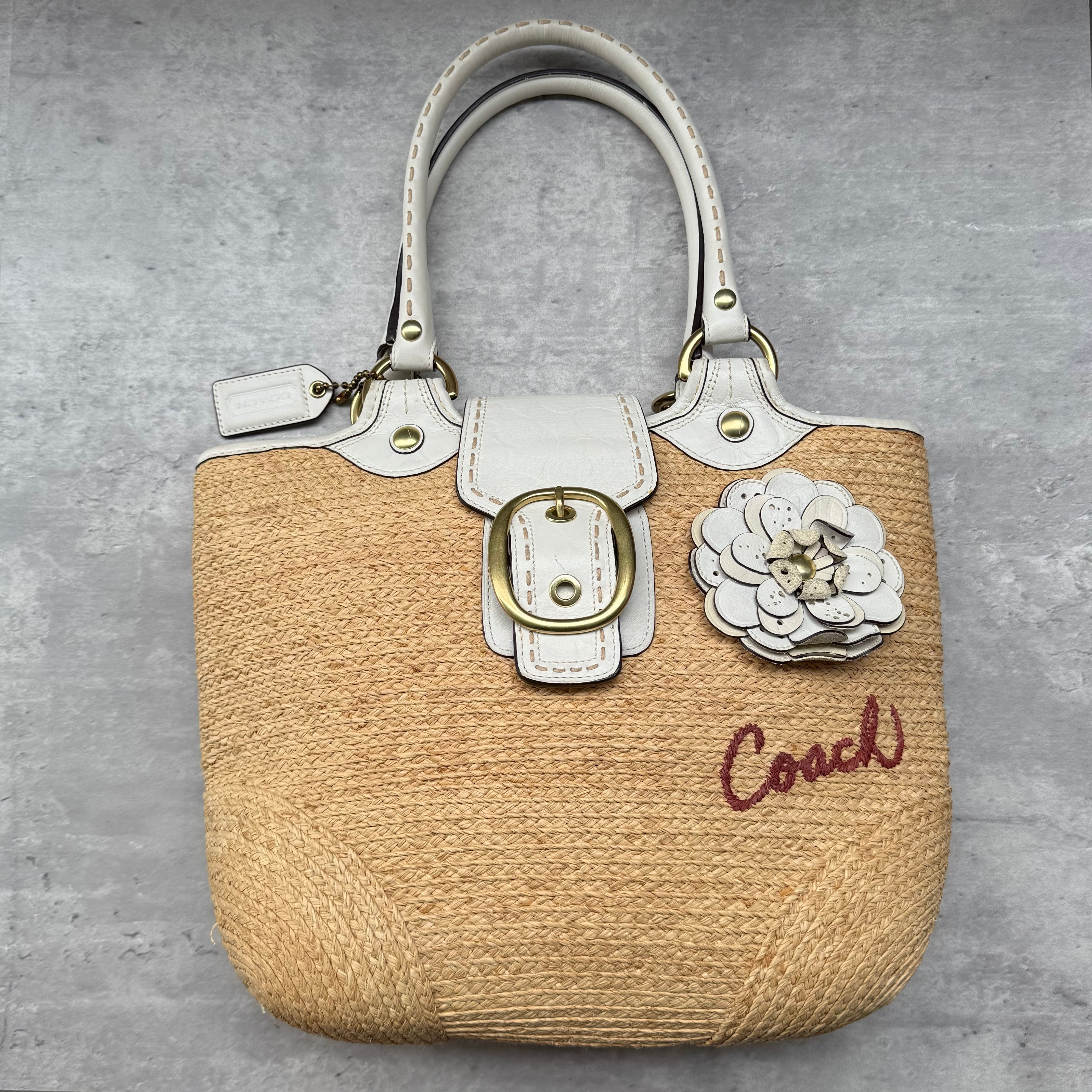 Coach Bleecker Straw Tote Bag in tan woven straw with white leather trim, floral appliqué, and multicolor check lining.