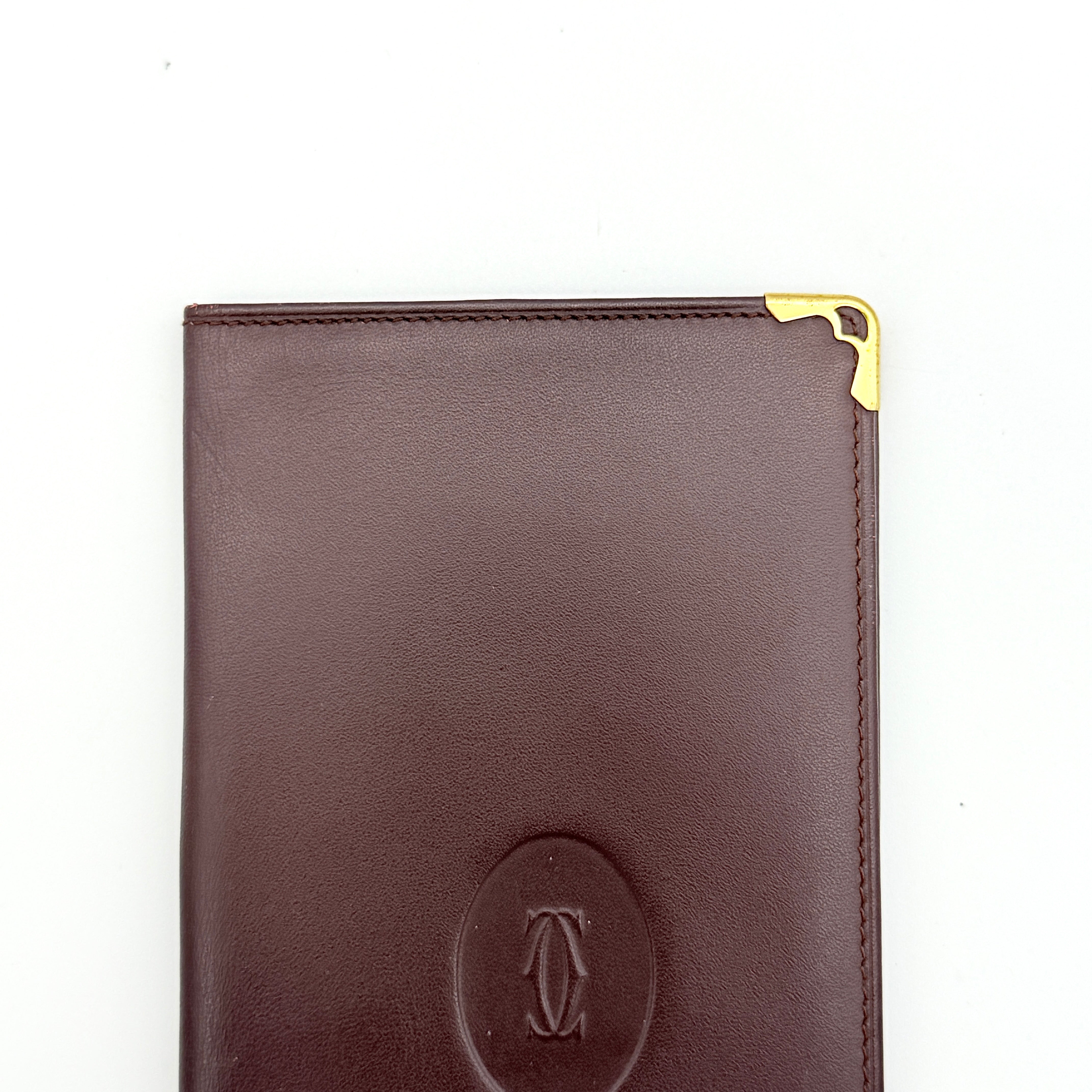 Cartier Must de Cartier bifold long wallet in burgundy leather with gold-tone corner accents, shown closed against a white background.
