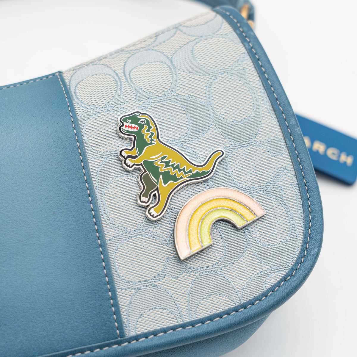 Coach 2Way Signature Swinger Shoulder Bag in light blue monogram canvas and leather with dinosaur and rainbow patches, shown with detachable strap.