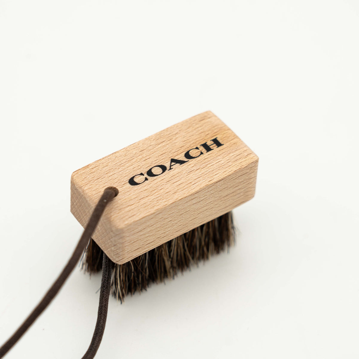Coach horsehair leather care brush with wood handle and brown bristles, featuring a Coach logo and leather cord loop.